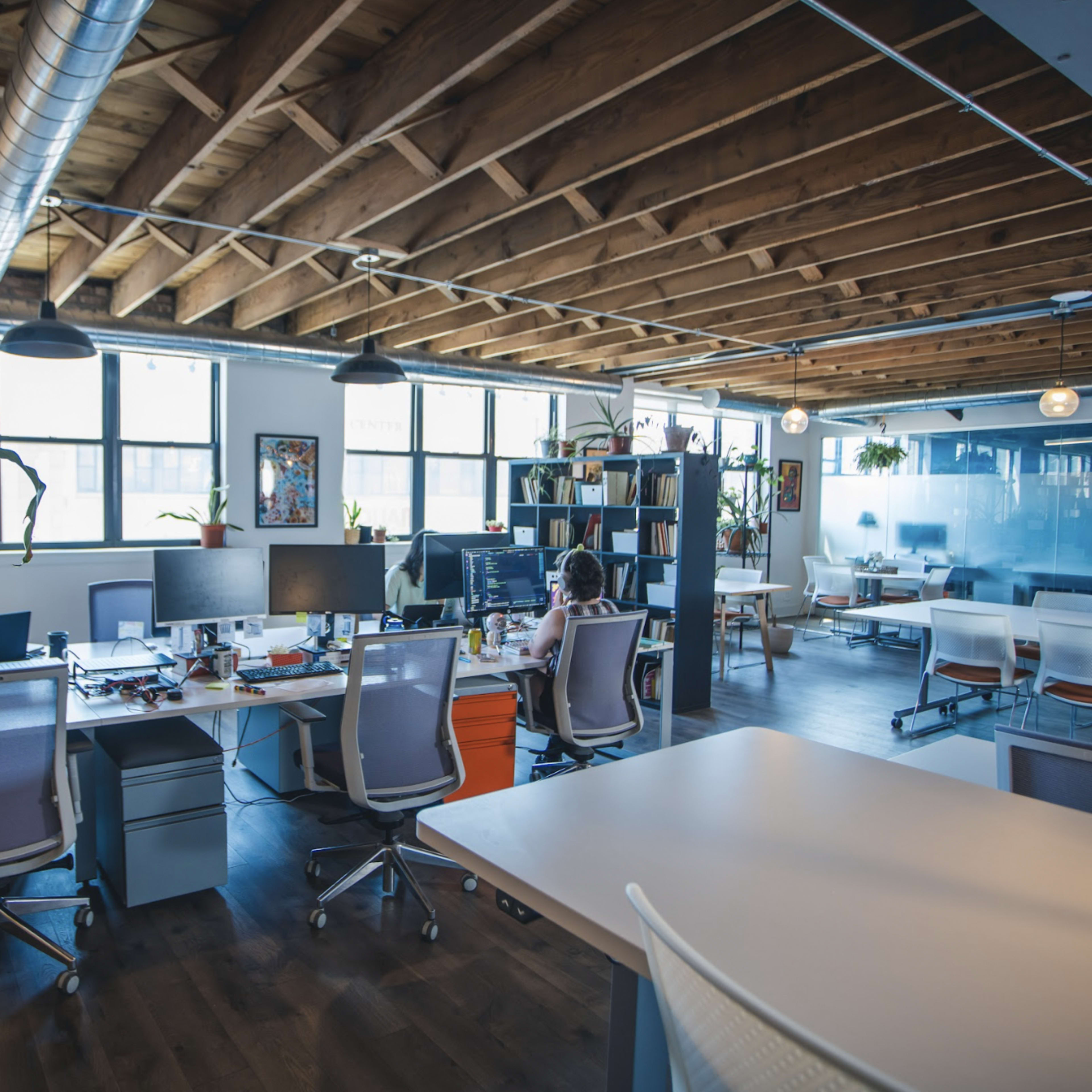 An open office space features several workstations equipped with computers and plants, with one person working at a desk in the background.