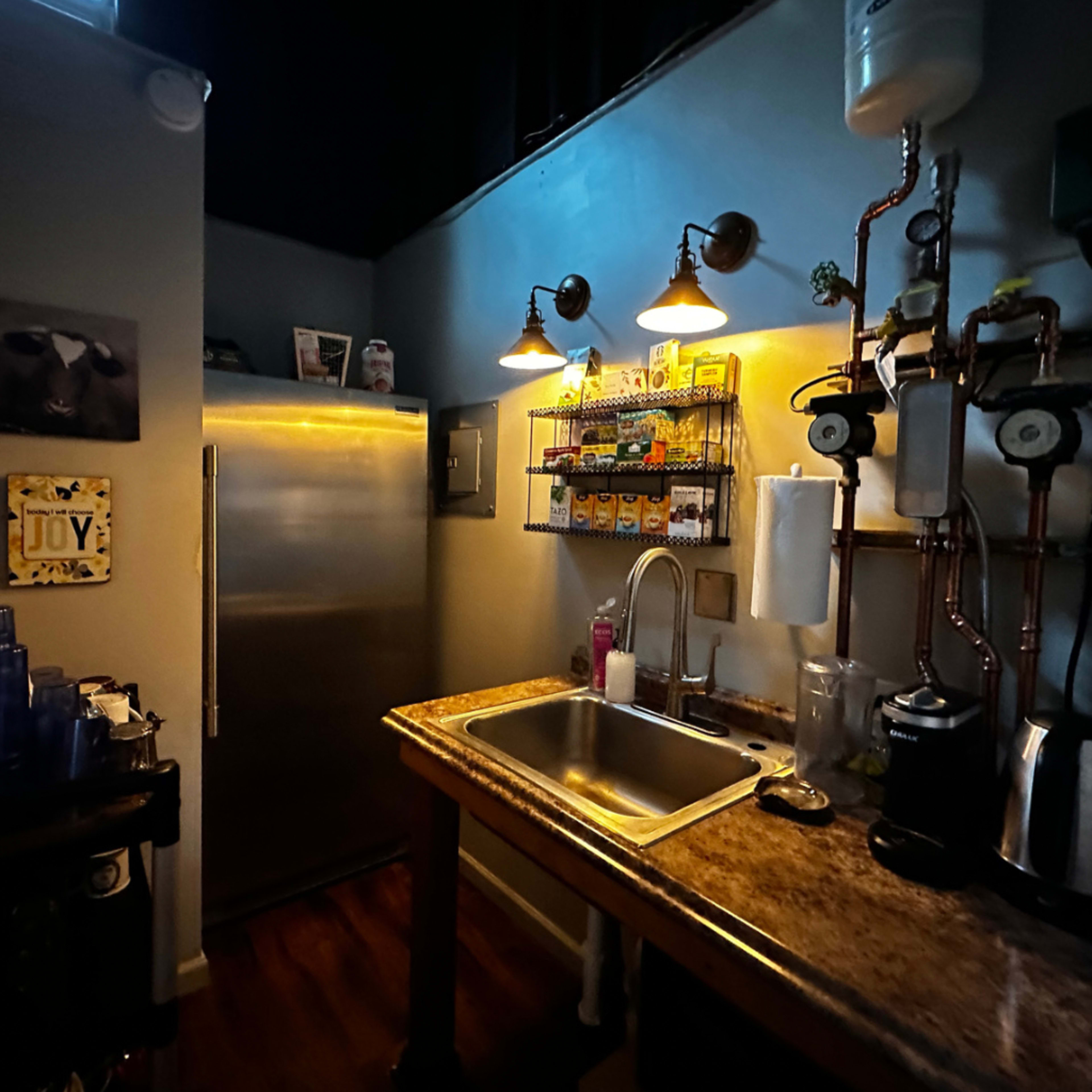A dimly lit kitchen area with a stainless steel refrigerator, a countertop sink, and shelves lined with various jars and containers.