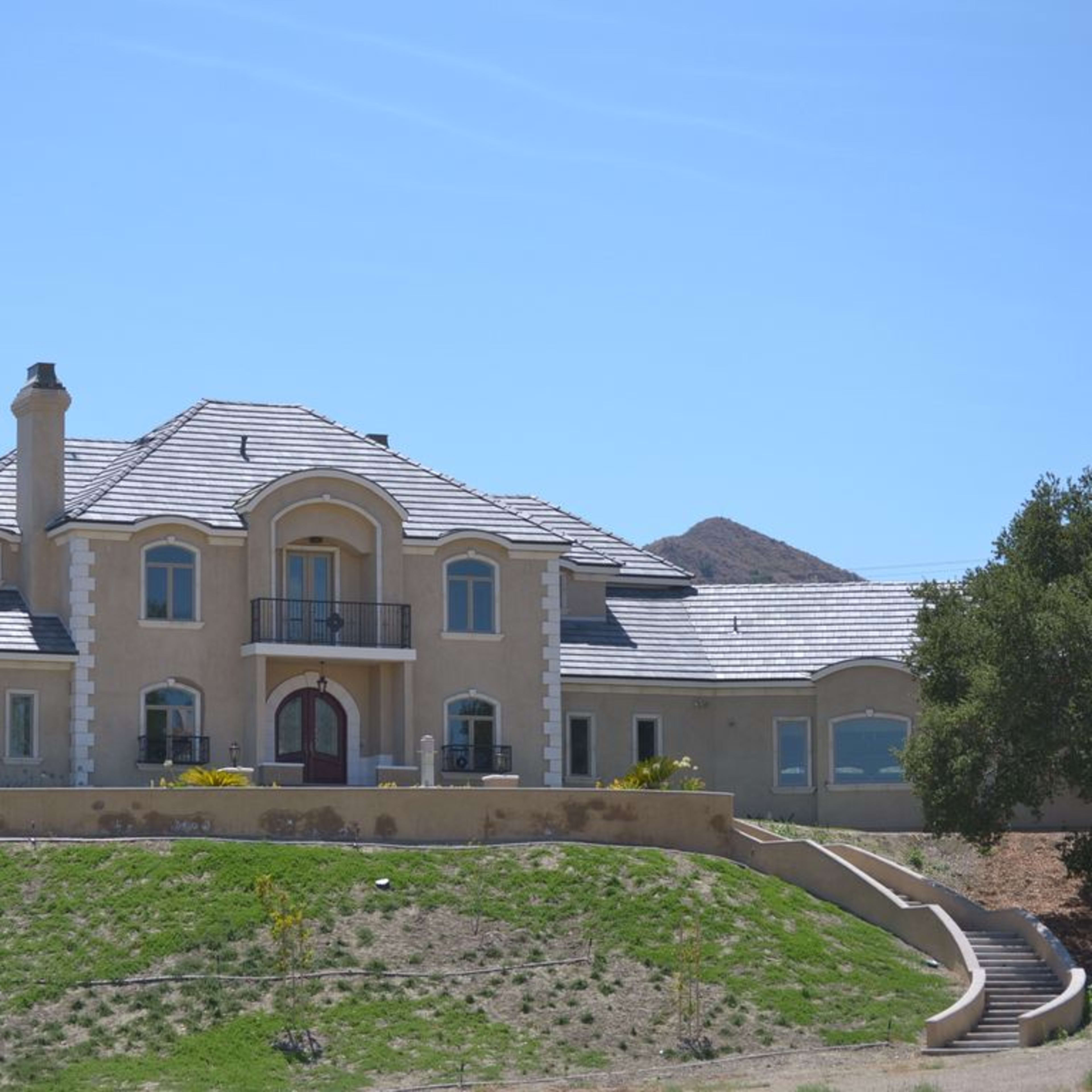 A large, two-story house with a tiled roof is situated on a grassy slope, surrounded by mountains.