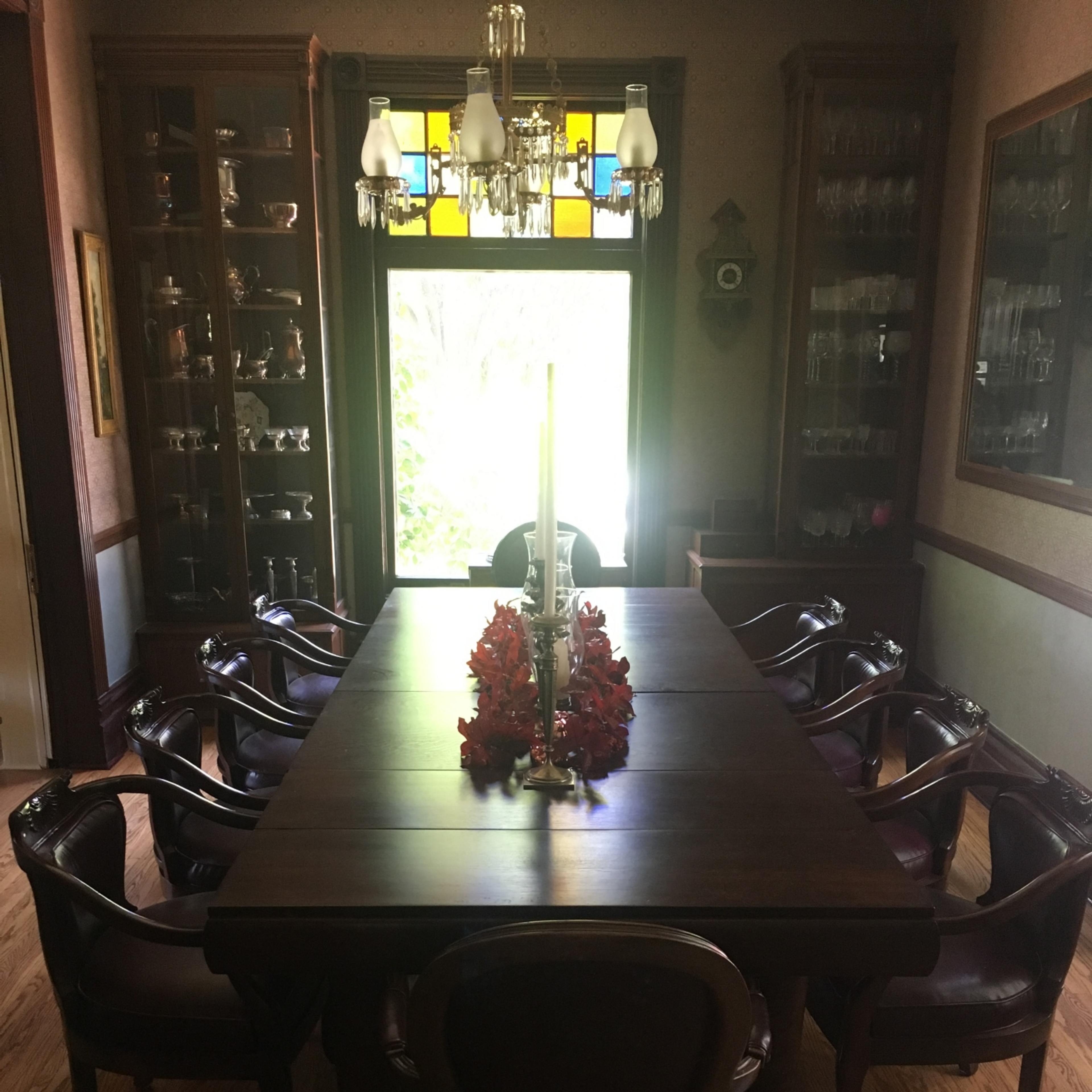 A long wooden dining table with eight chairs is set in a well-lit room featuring stained glass windows and a floral centerpiece.