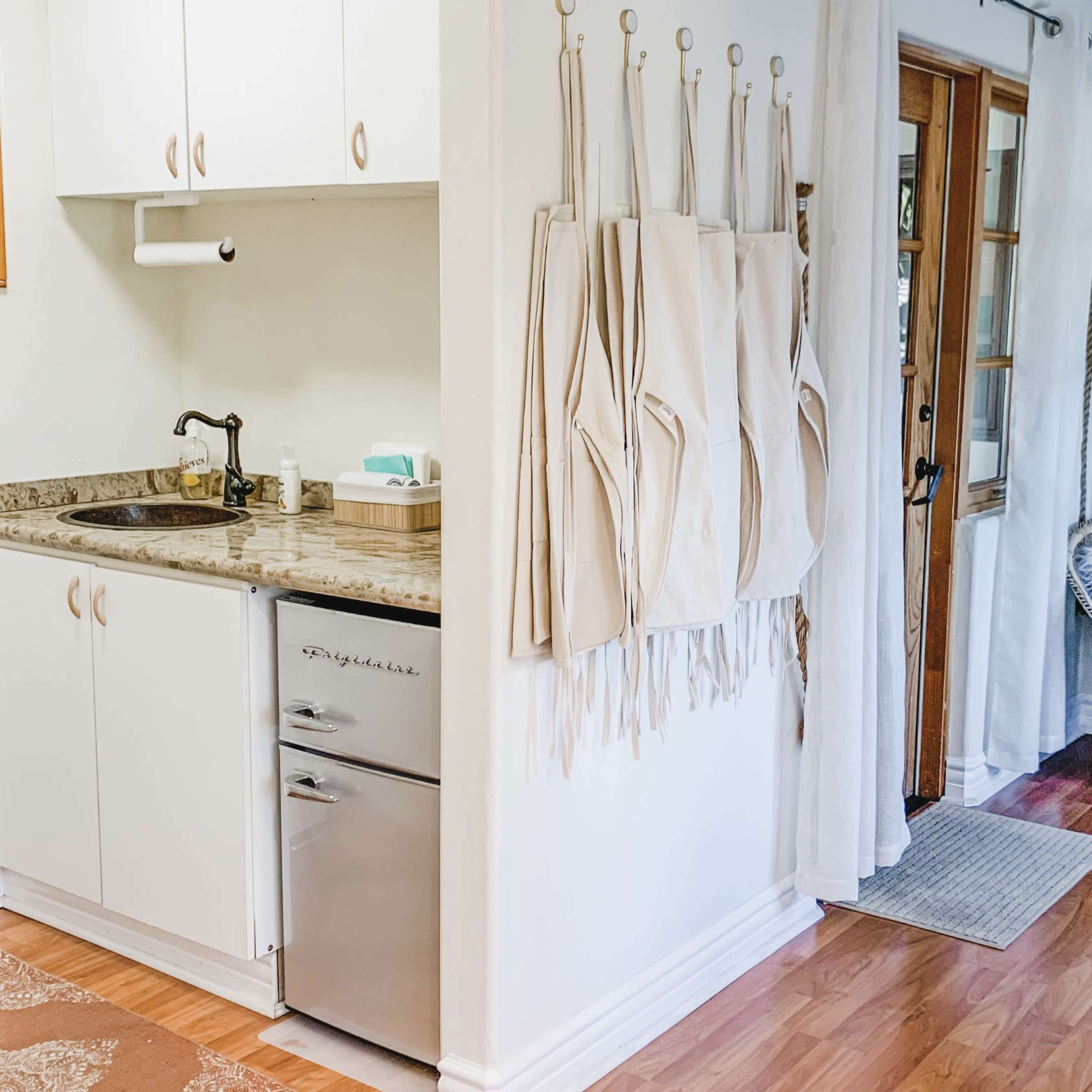 A kitchen area with a sink, a small refrigerator, and several beige aprons hanging on the wall.