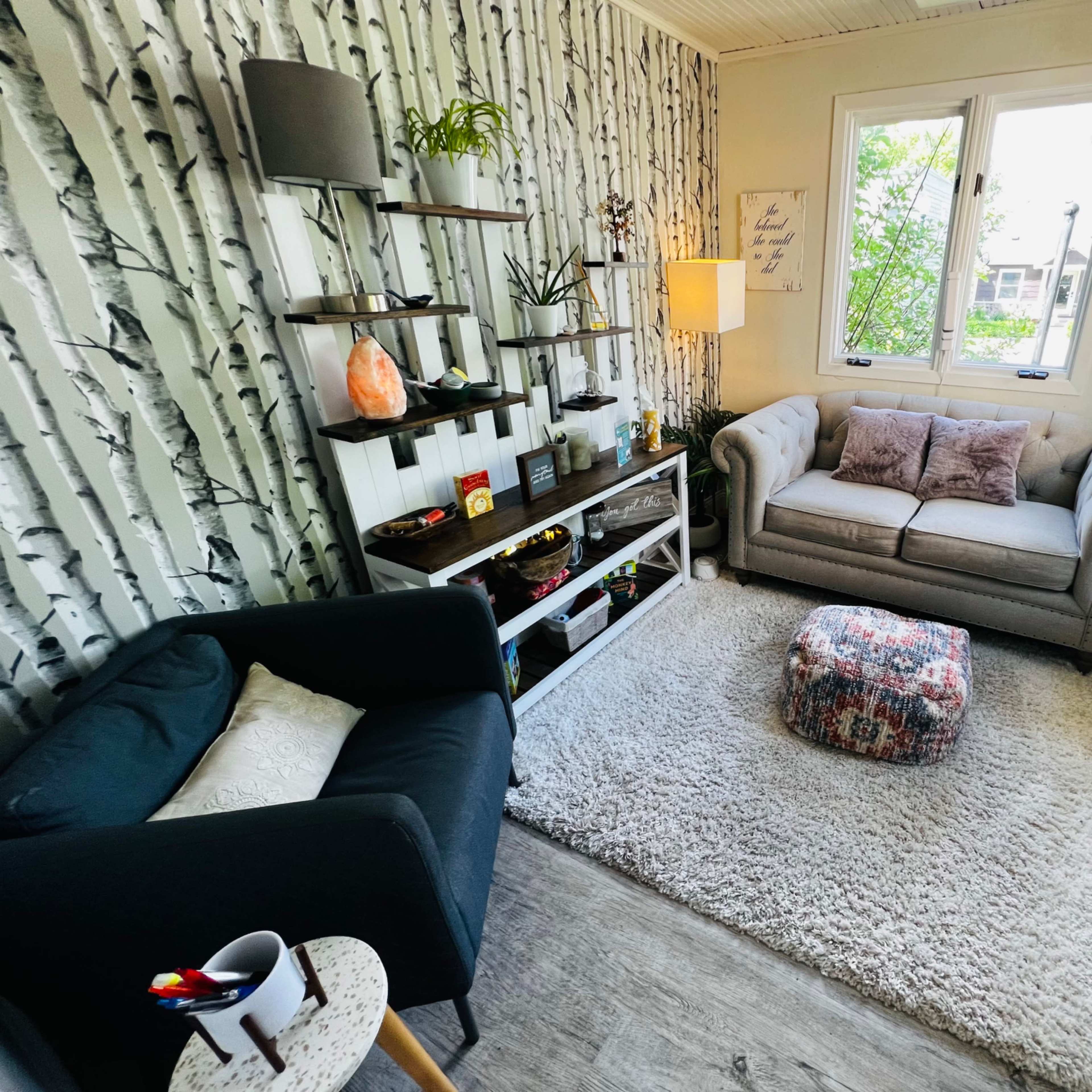 A cozy room featuring a gray sofa, a black chair, a patterned ottoman, and shelves with decorative items against a birch tree-patterned wall.