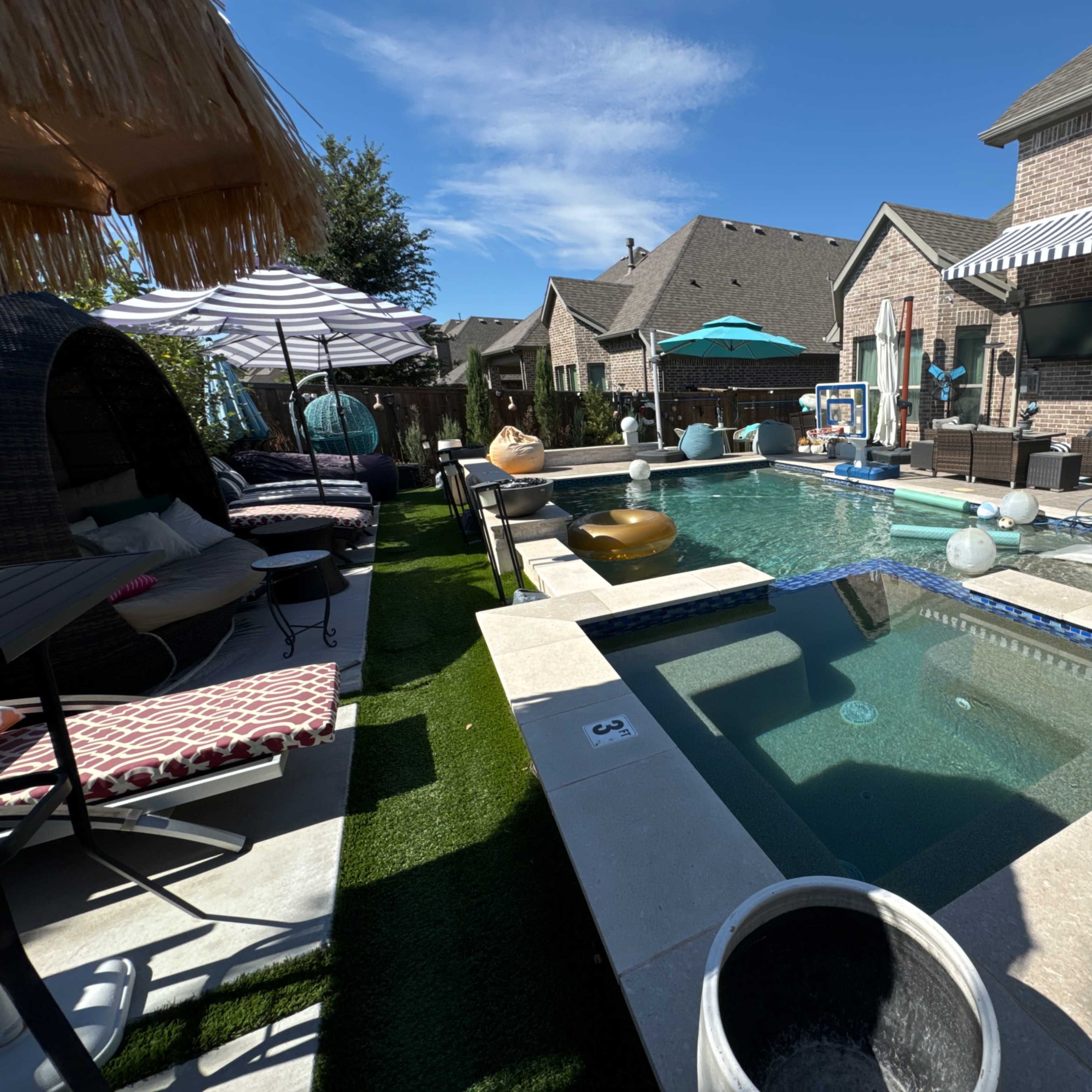 A residential backyard featuring a swimming pool with a hot tub, surrounded by lounge chairs and shaded seating areas.