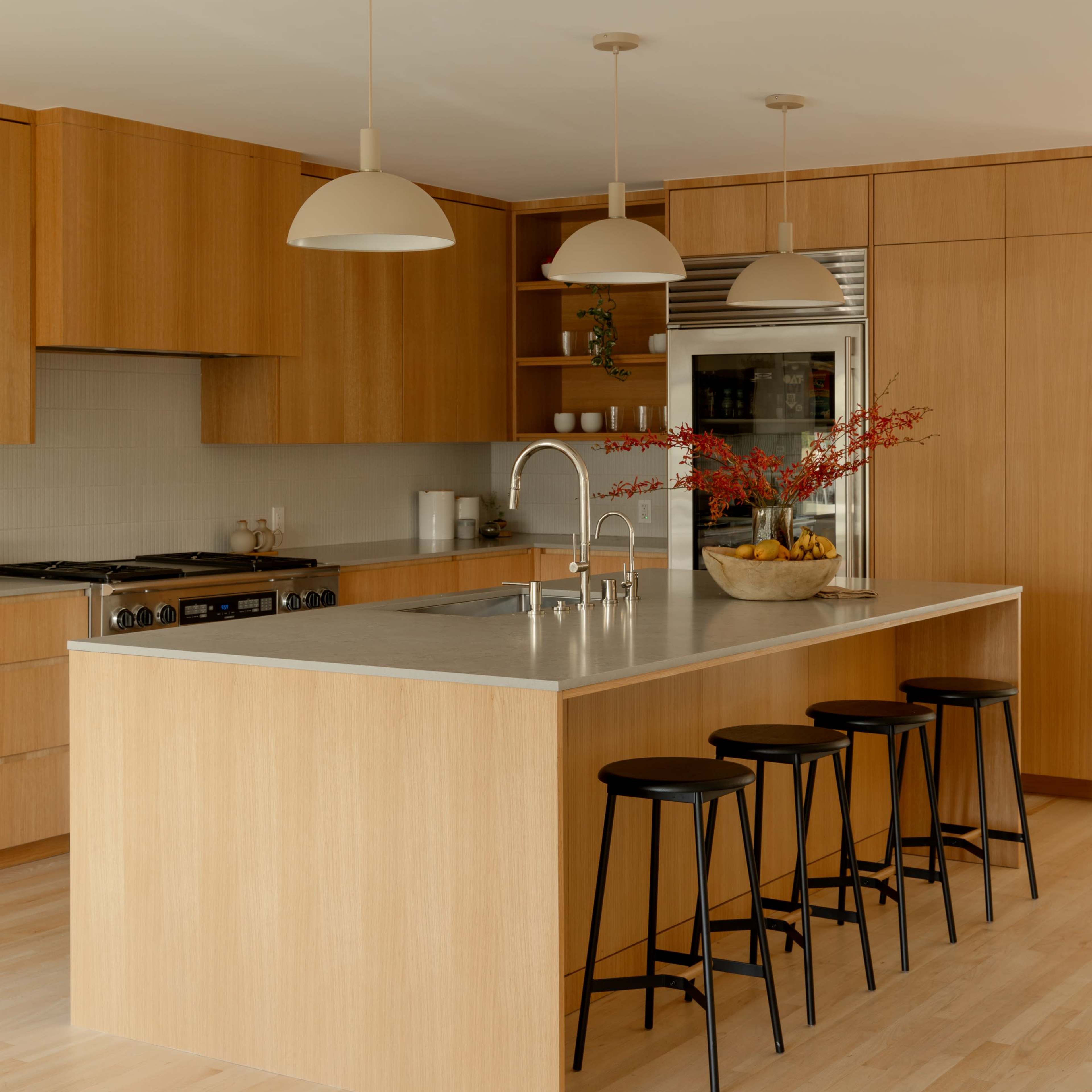 A modern kitchen features wooden cabinetry, a large island with a stainless steel countertop, and four black stools.