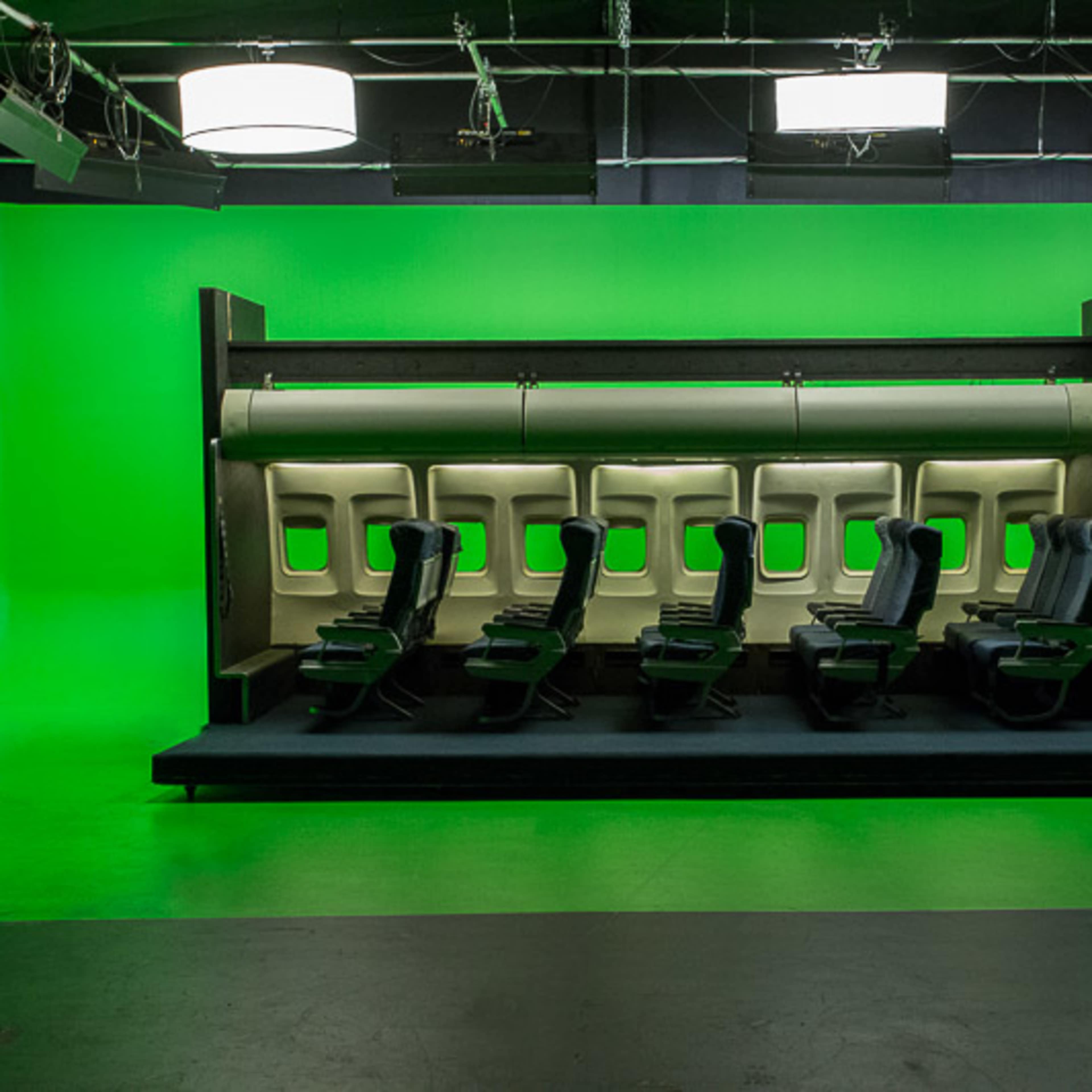 A row of airplane seats set against a bright green screen backdrop in a studio environment.