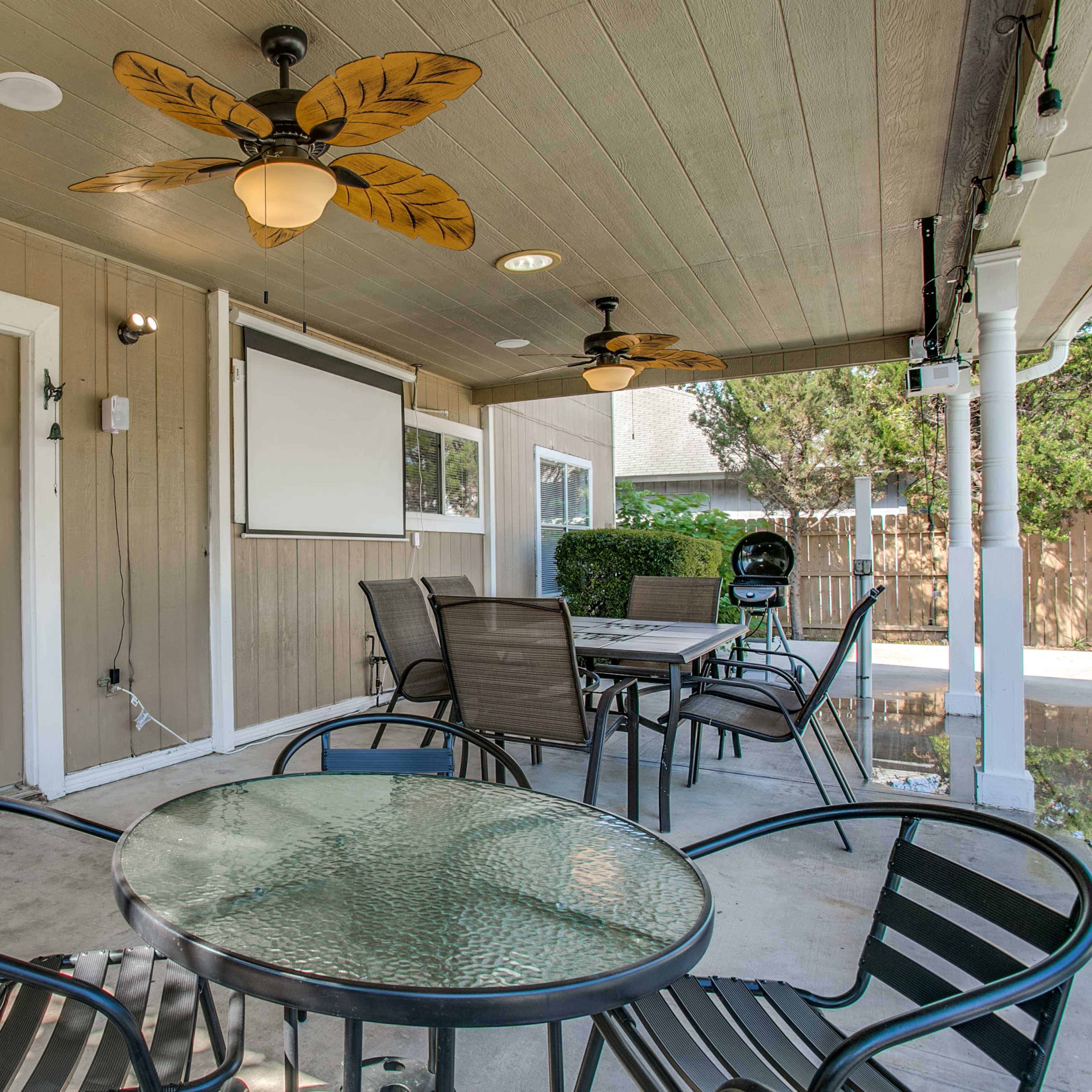 A covered outdoor patio with a glass table and several chairs, featuring a ceiling fan and a wall-mounted screen.