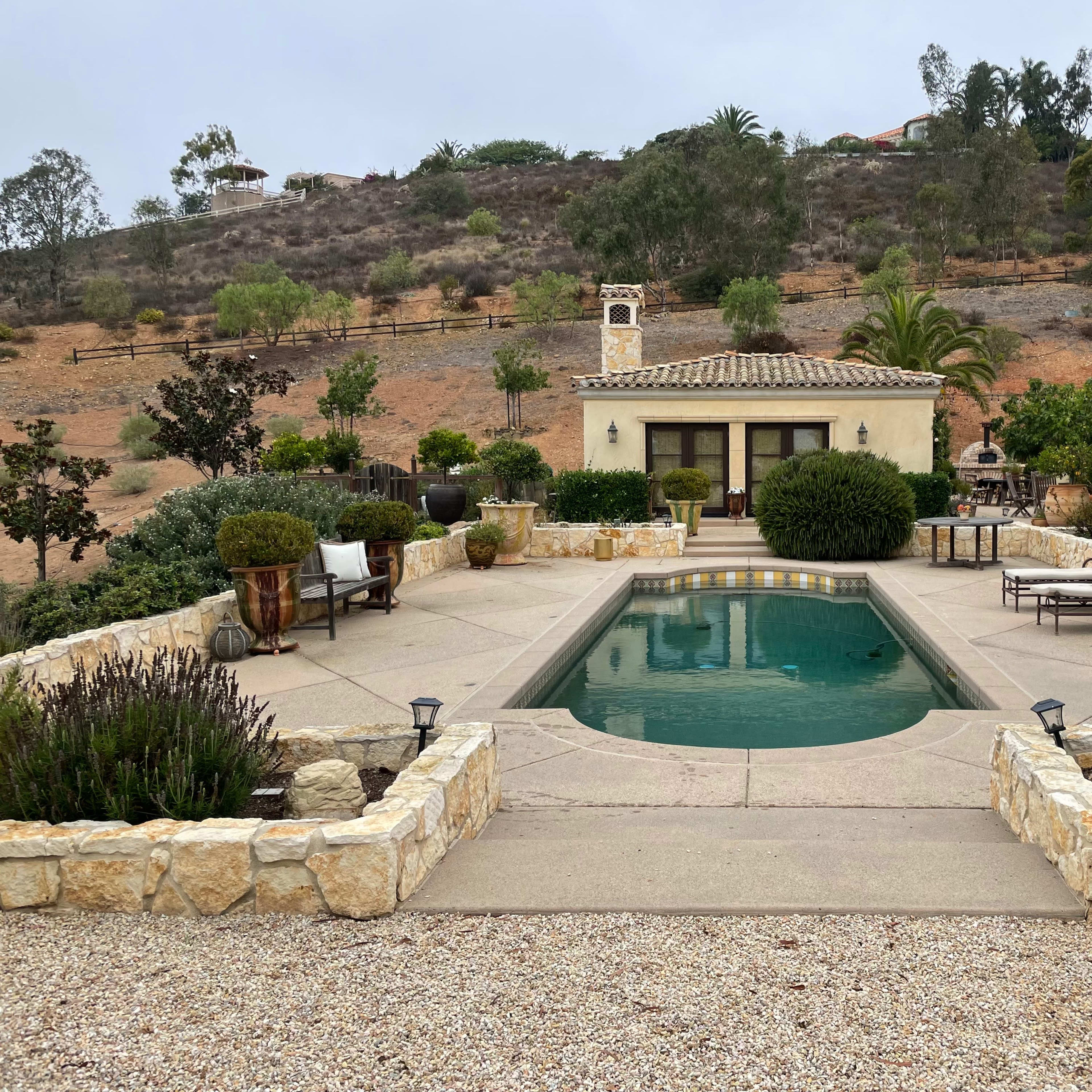 A landscaped outdoor area featuring a swimming pool surrounded by stone patios, lounge chairs, and various plants, set against a hillside.