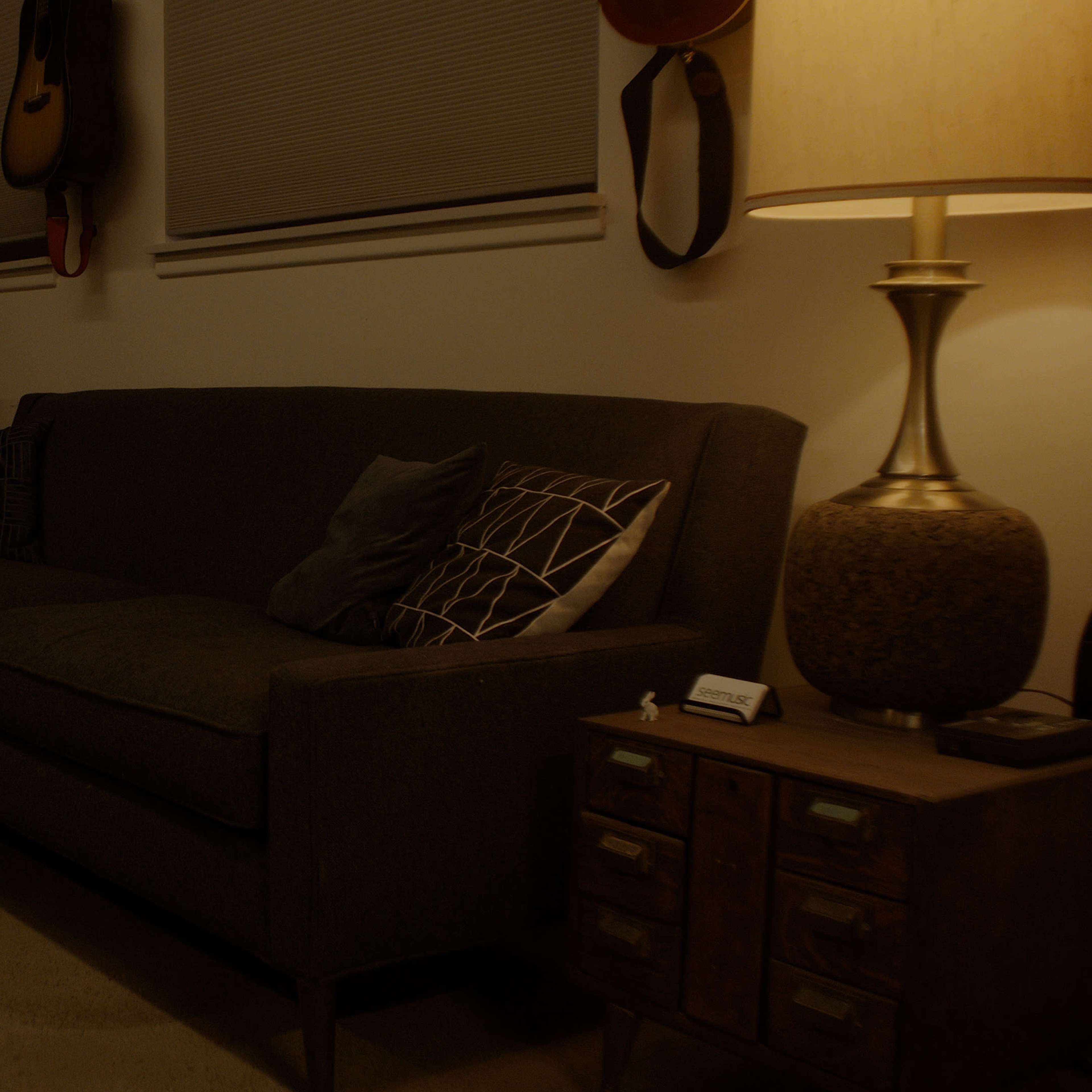 A dimly lit living room with a dark sofa, a small table, a lamp, and a keyboard in the corner.