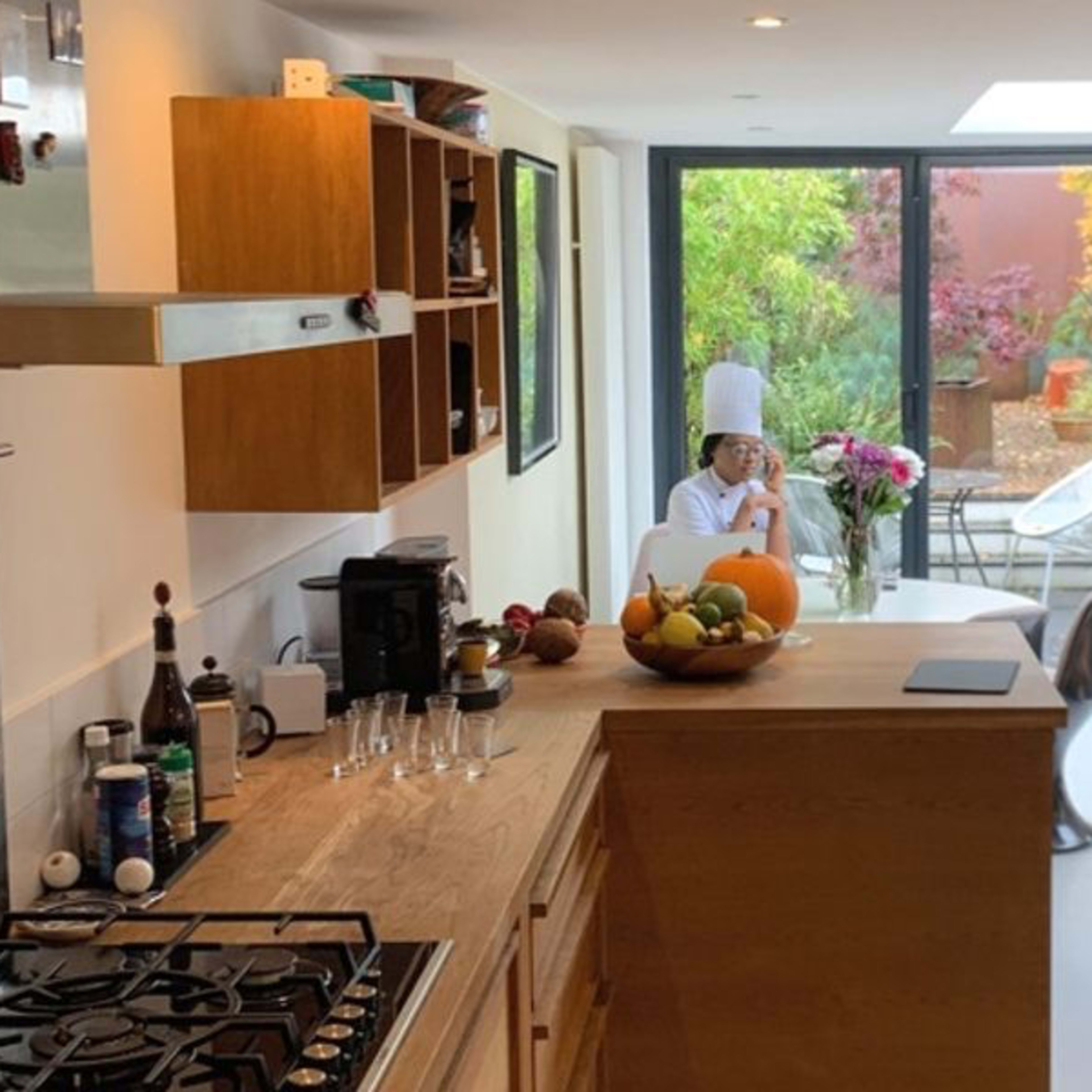 A modern kitchen opens up to a bright dining area with a view of a garden, featuring a person seated at a table surrounded by plants and natural light.
