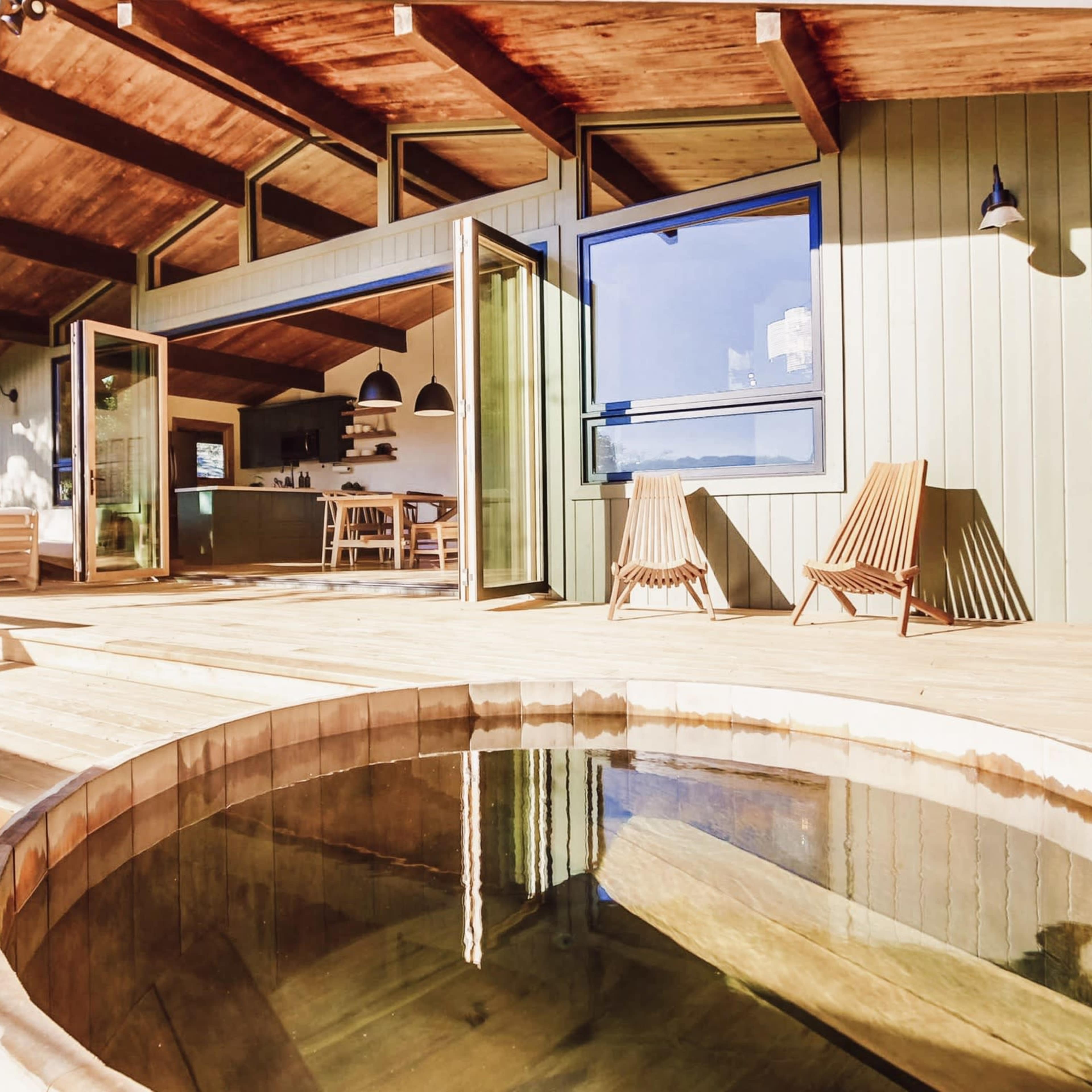 A wooden deck features an outdoor hot tub with two wooden lounge chairs and expansive views of the surrounding trees.