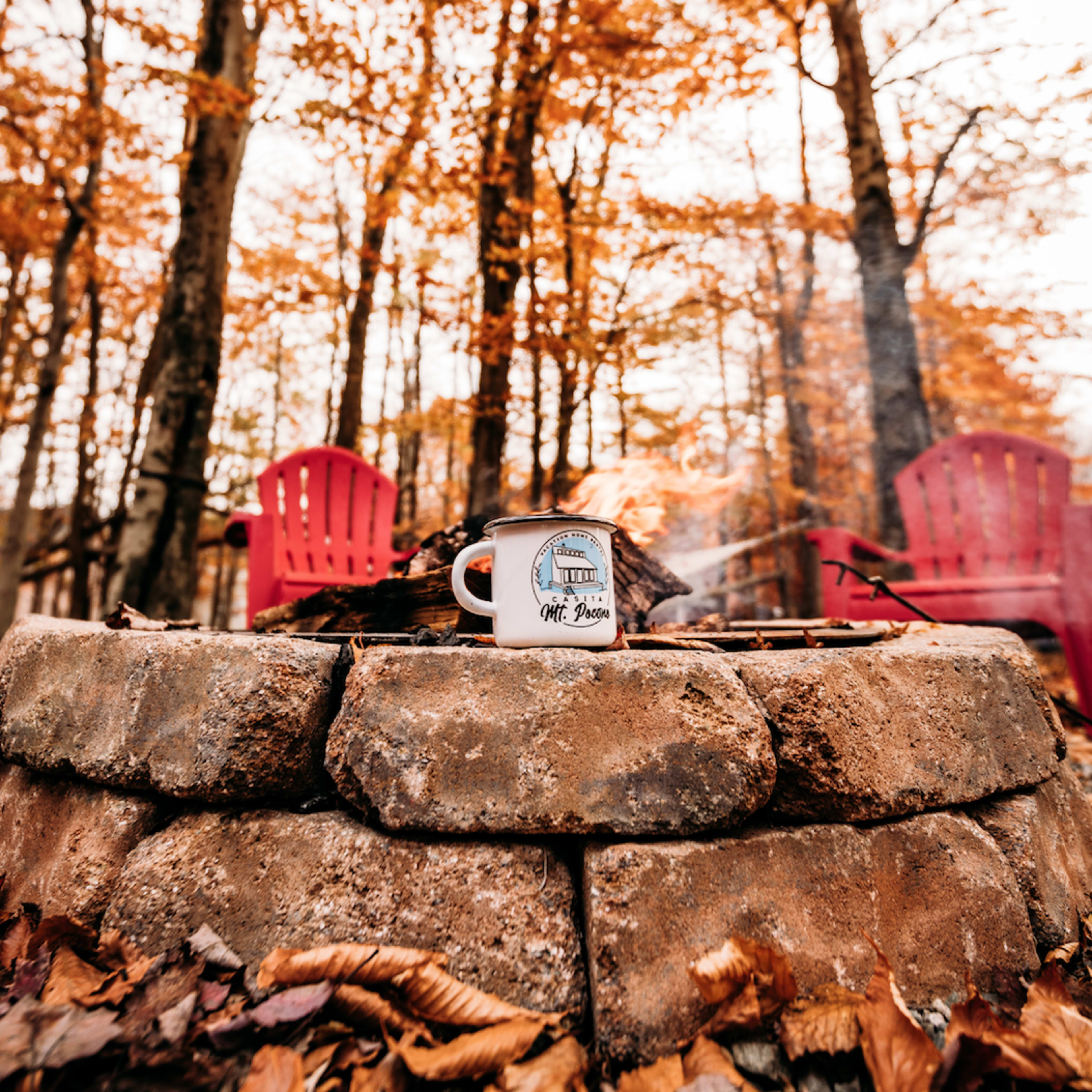 A mug sits on a stone fire pit surrounded by red chairs and autumn leaves under a canopy of orange trees.