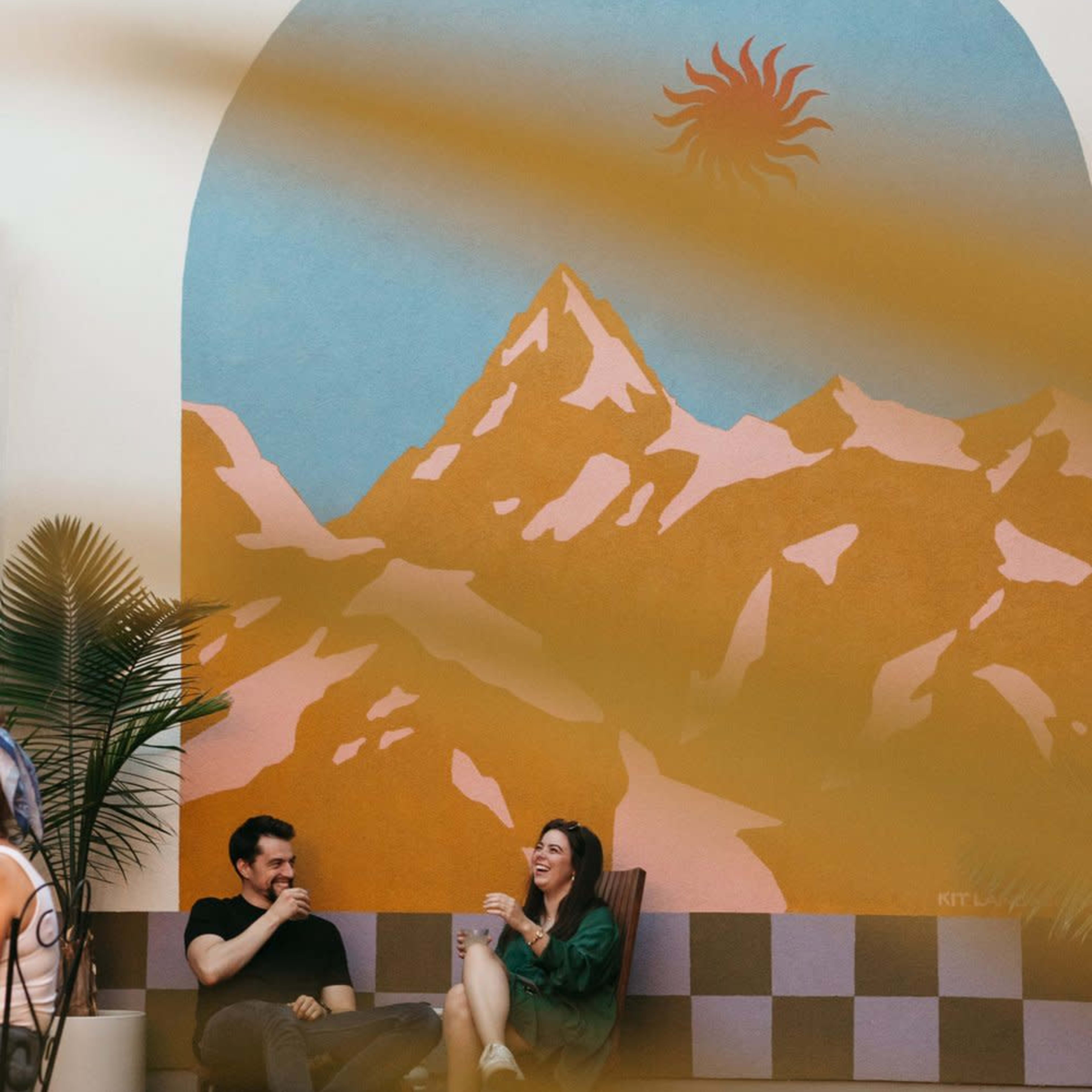 Two people sit on chairs in front of a colorful mural depicting mountains and a sun.