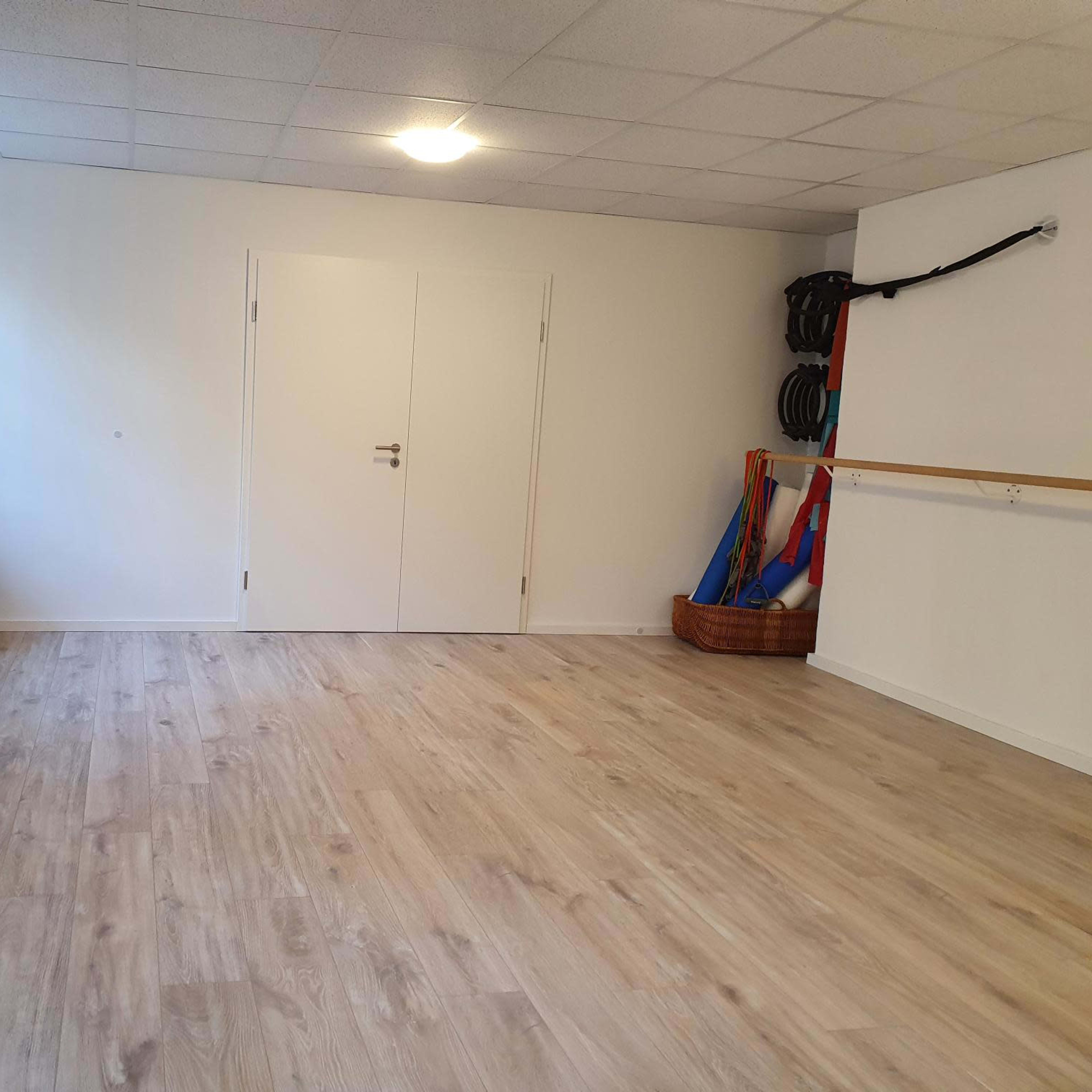A spacious, empty room with wooden flooring, a large window, and exercise equipment stored against the wall.