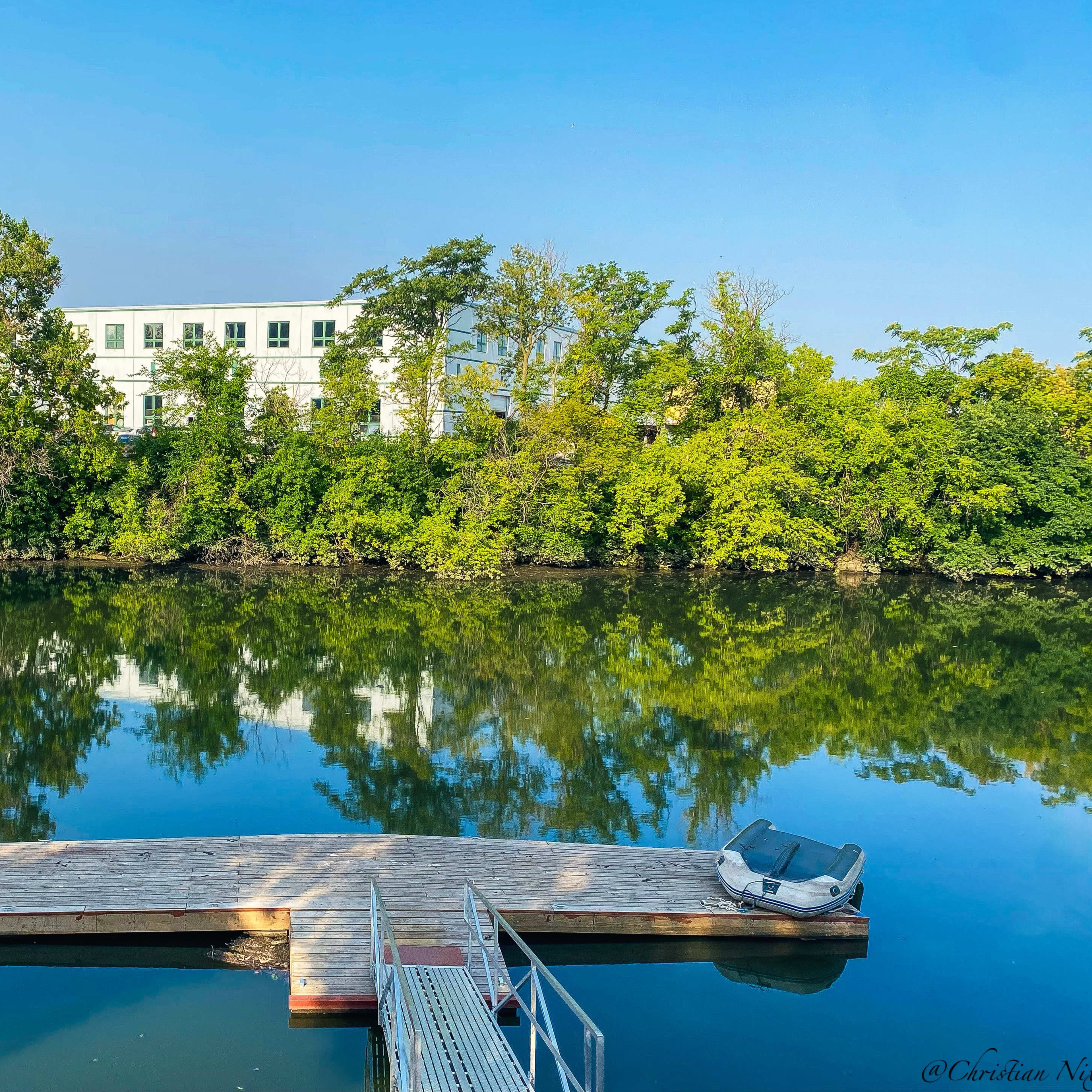 A wooden dock extends over a calm river, reflecting trees and a building on the opposite bank.
