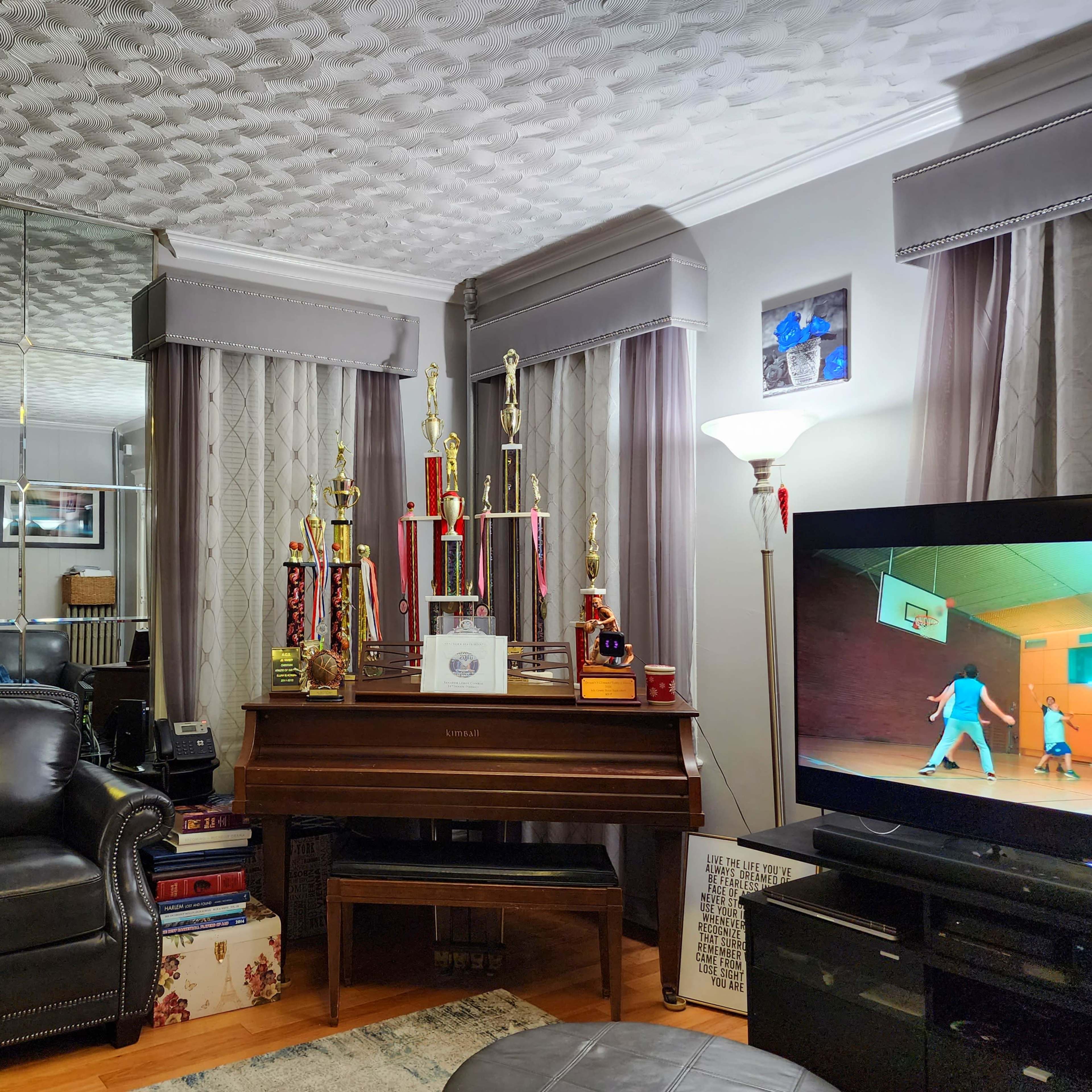 A living room with a black leather chair, a grand piano adorned with trophies, and a television displaying a basketball game scene.