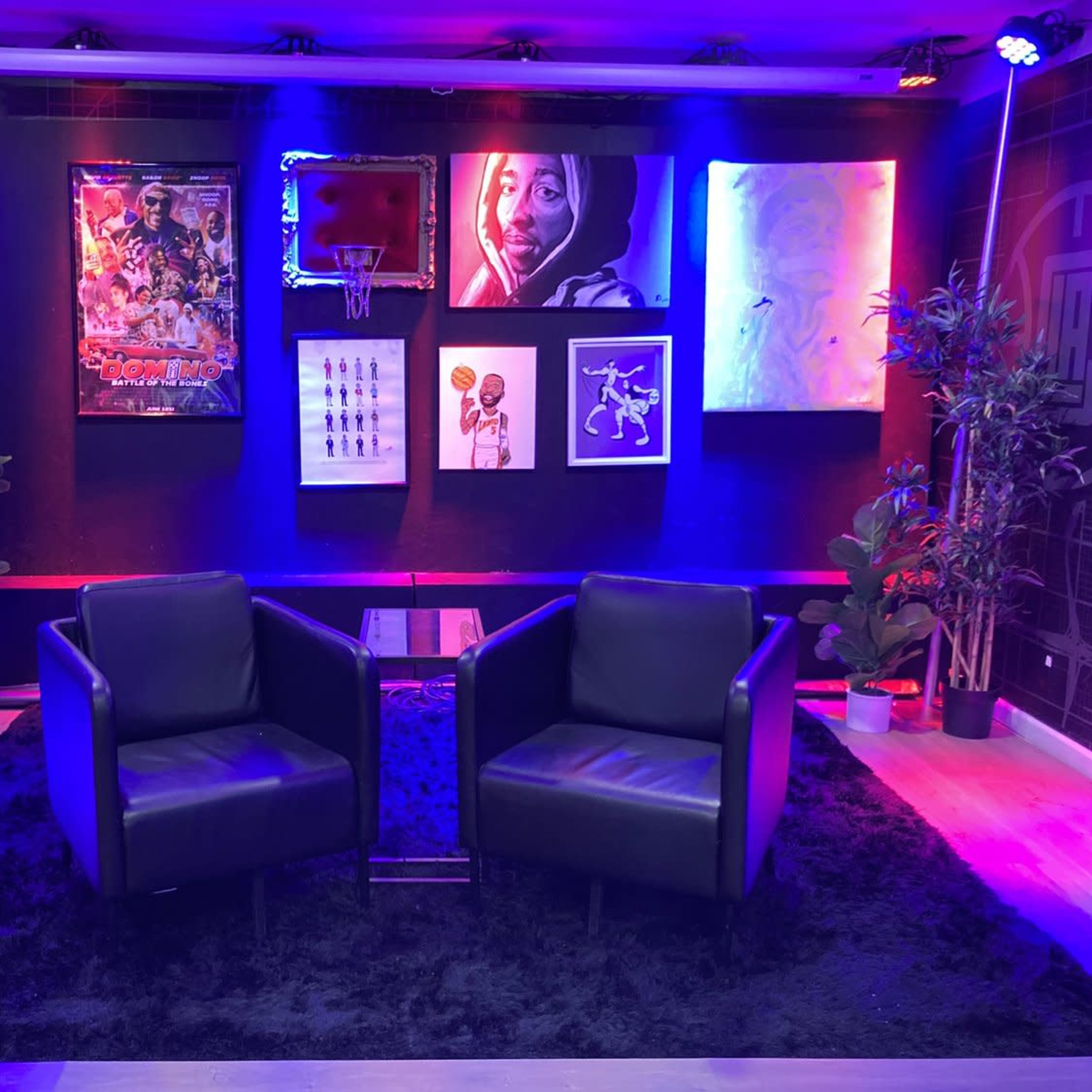 A modern seating area with two black chairs, a small glass table, and a gallery of framed artworks on the wall, illuminated by colorful lighting.