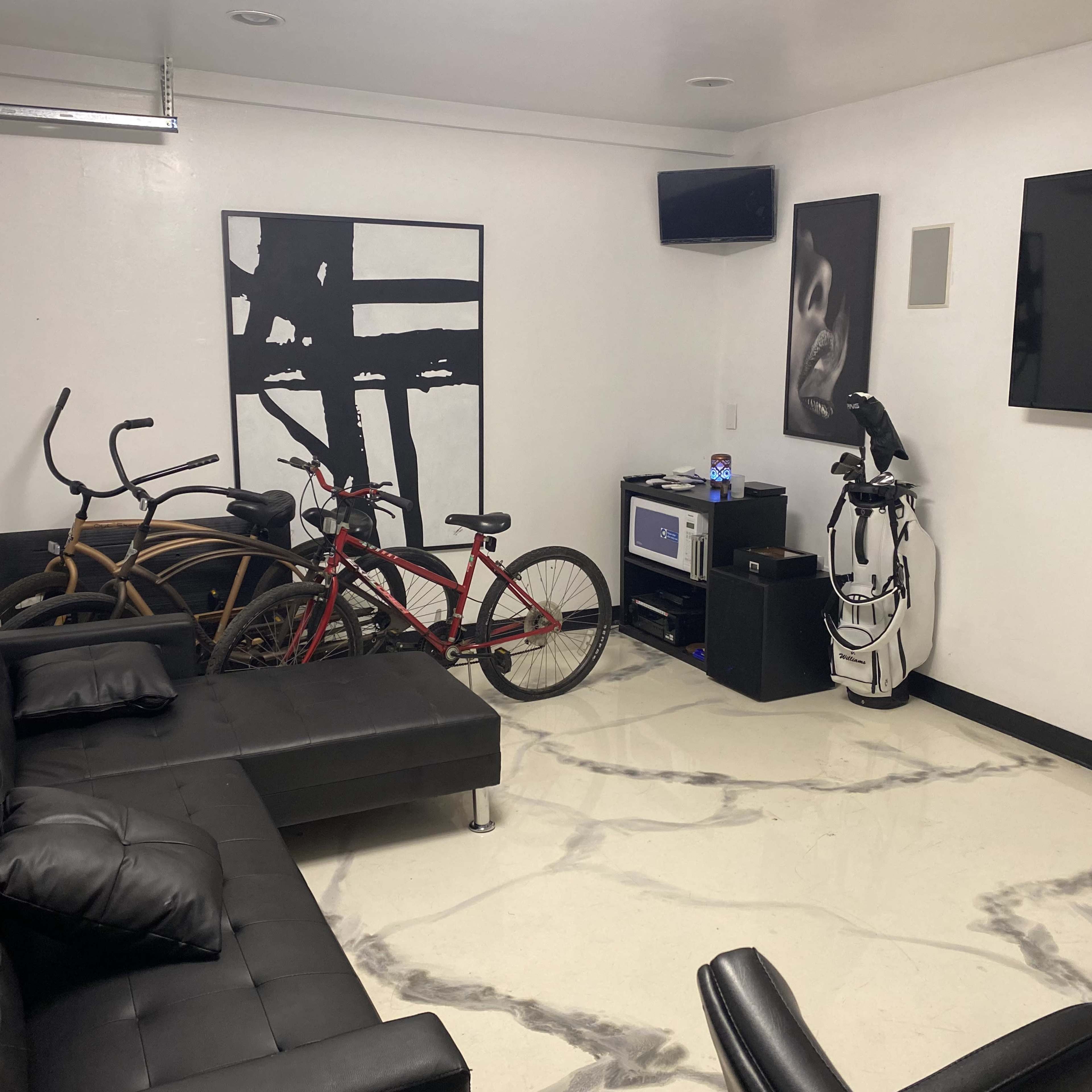 A modern room features two bicycles parked near a sleek black couch, a television on the wall, and minimalist artwork.