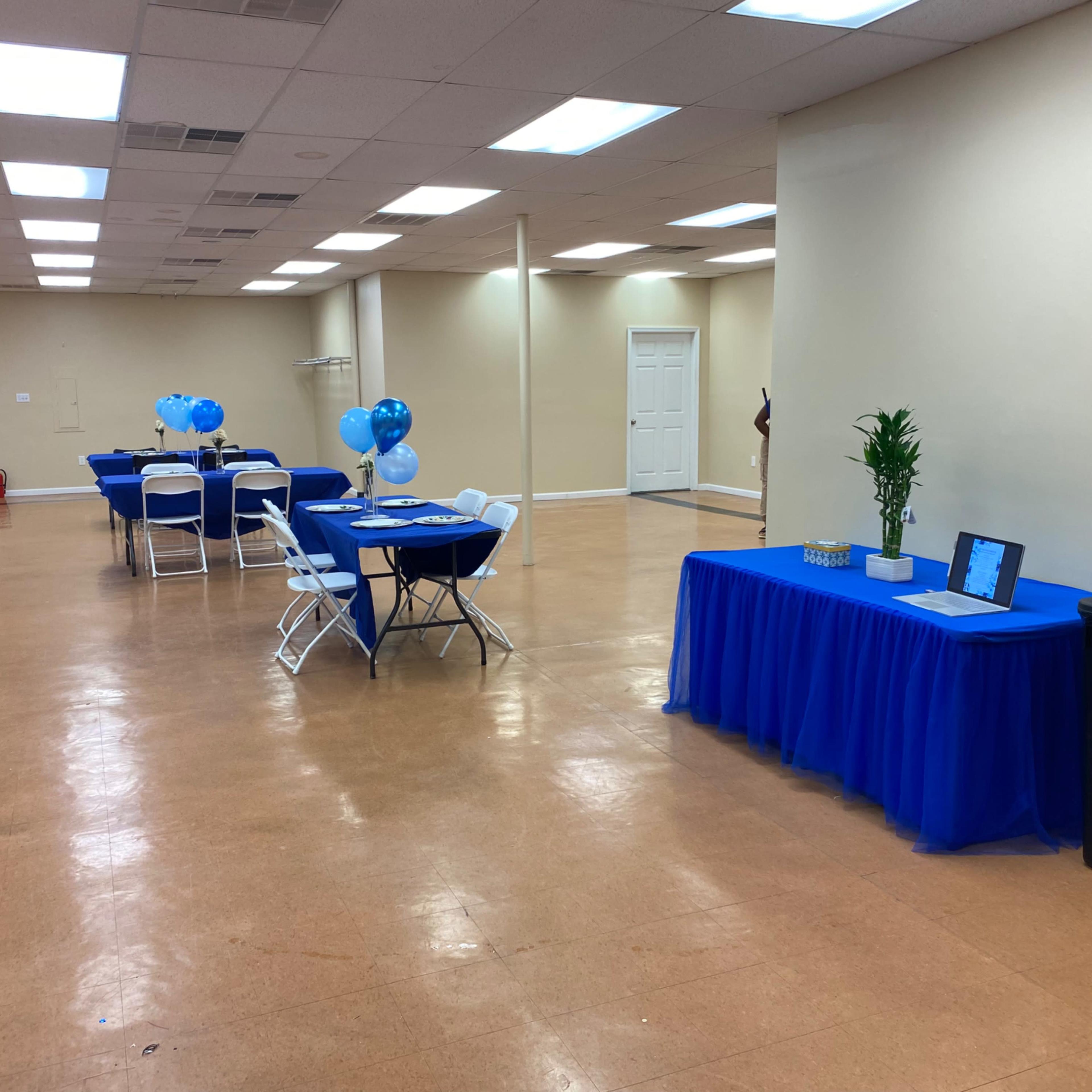 A large, empty room features several tables covered with blue tablecloths and balloons, along with a table displaying decorative items and a trash can.