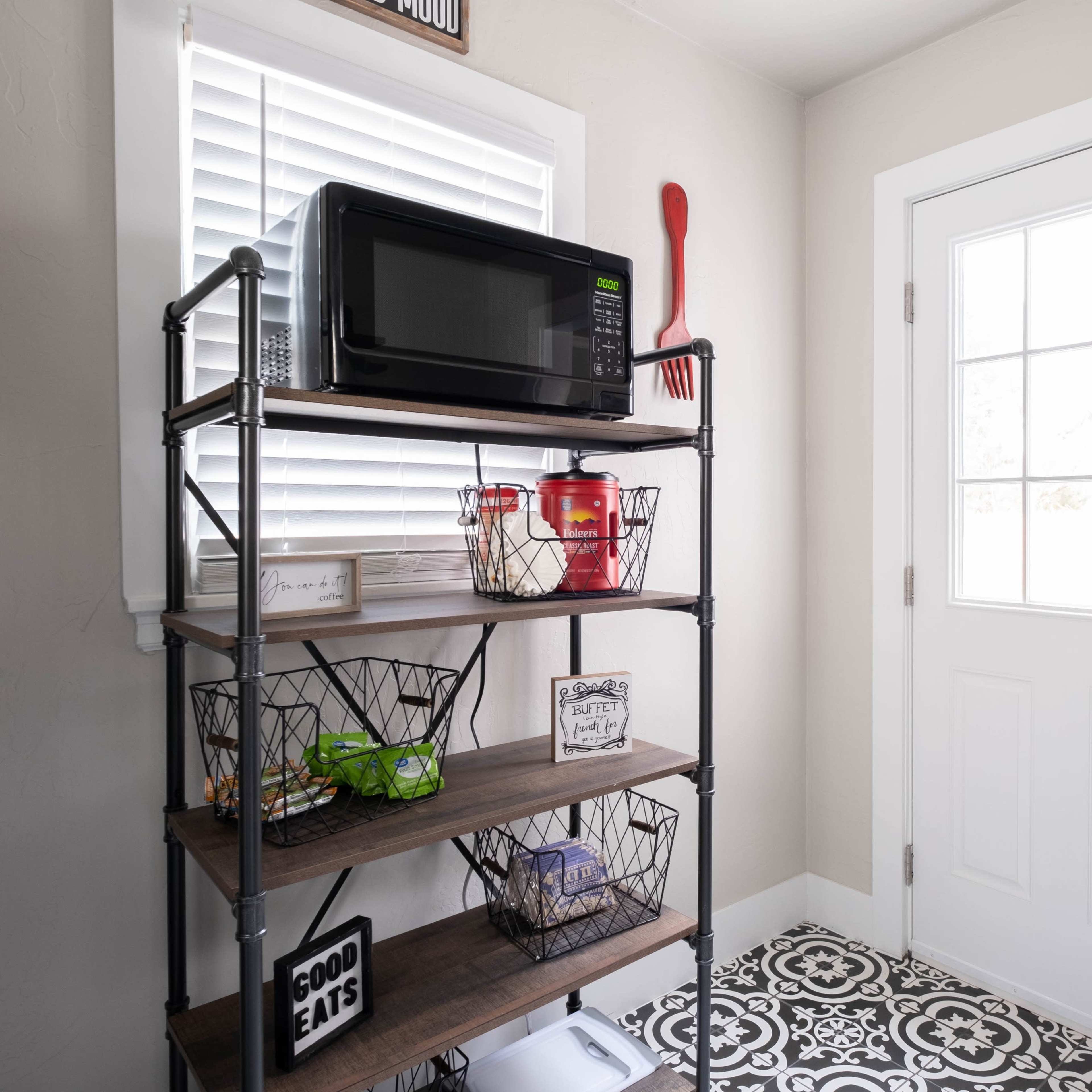 A metal shelving unit with a microwave, snacks in baskets, and decorative signs is positioned next to a door with glass panes and a window.