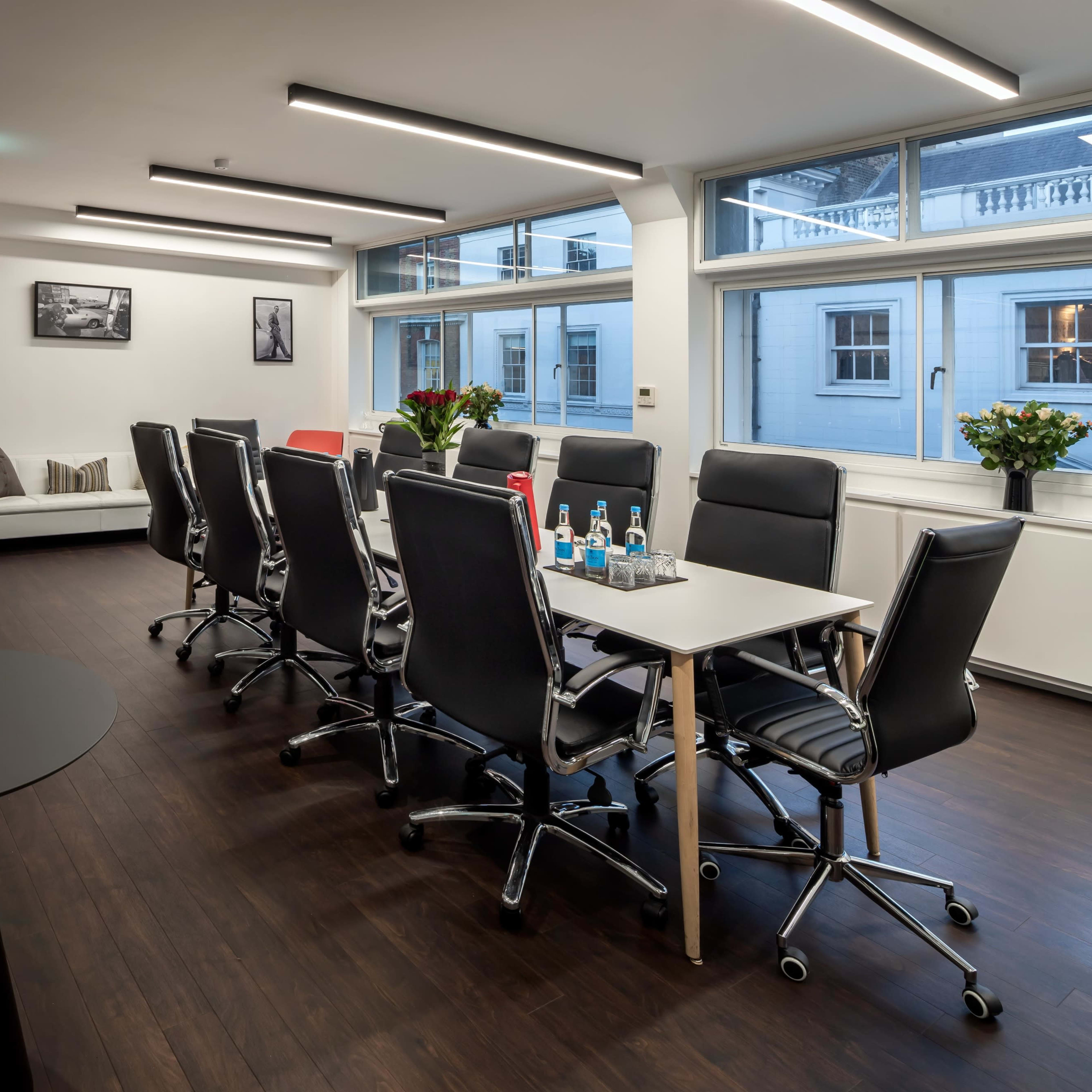 A modern conference room with a long table surrounded by black and red chairs, large windows providing natural light, and a few decorative elements like vases and artwork.