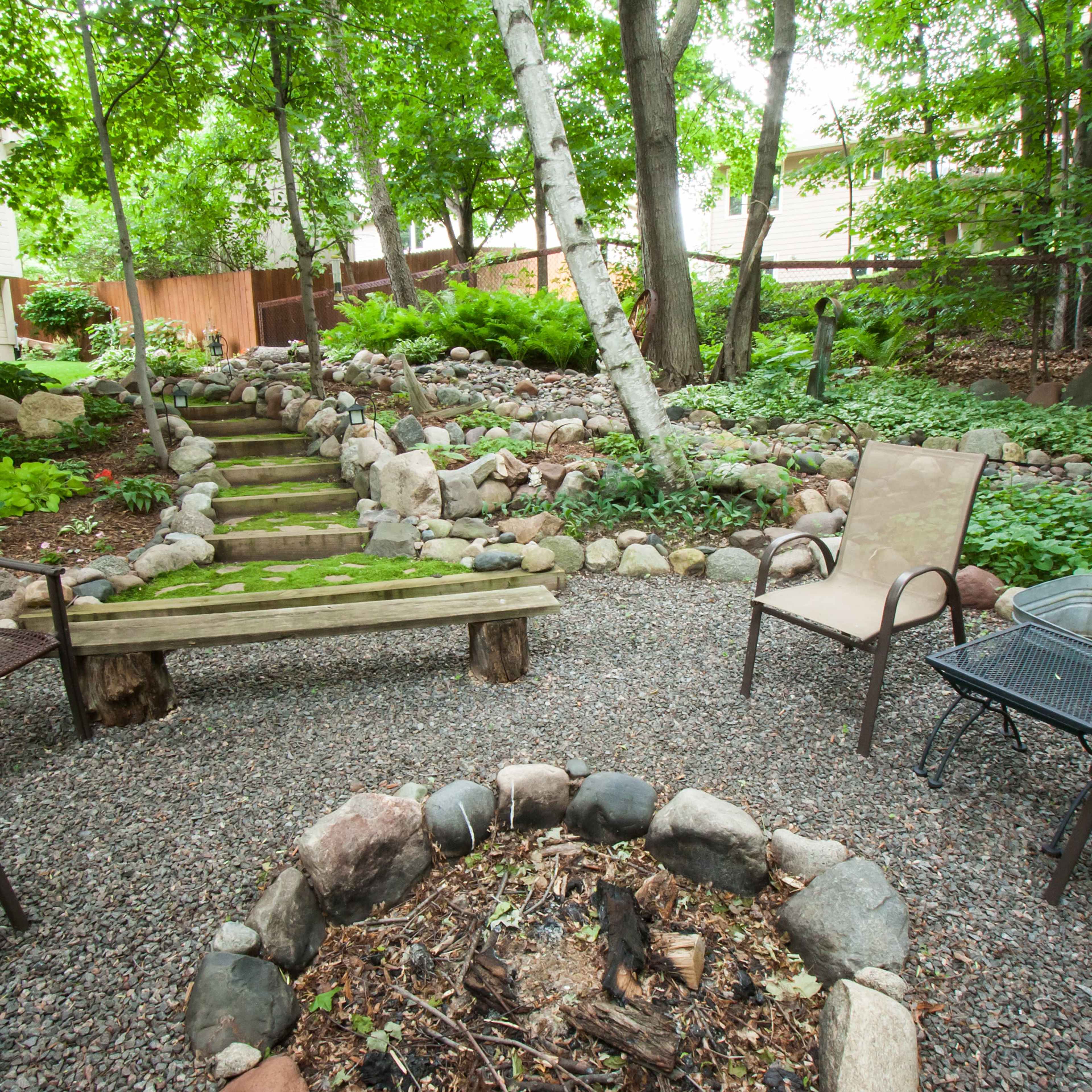 A landscaped backyard with a stone fire pit surrounded by chairs, a wooden bench, and a winding pathway through greenery and trees.