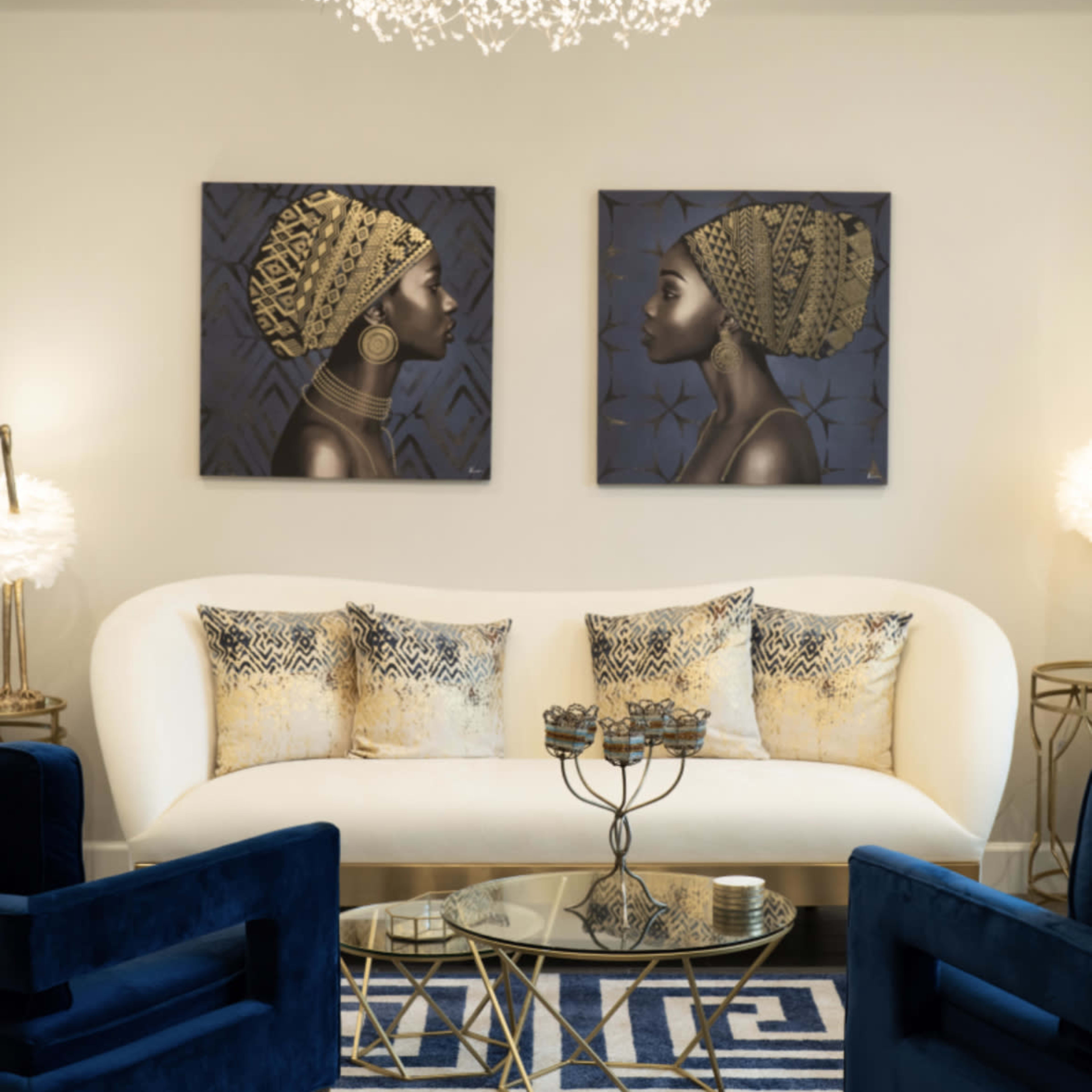 A modern living room featuring a white curved sofa with decorative pillows, two dark blue chairs, a round glass coffee table, and two framed artworks on the wall above.
