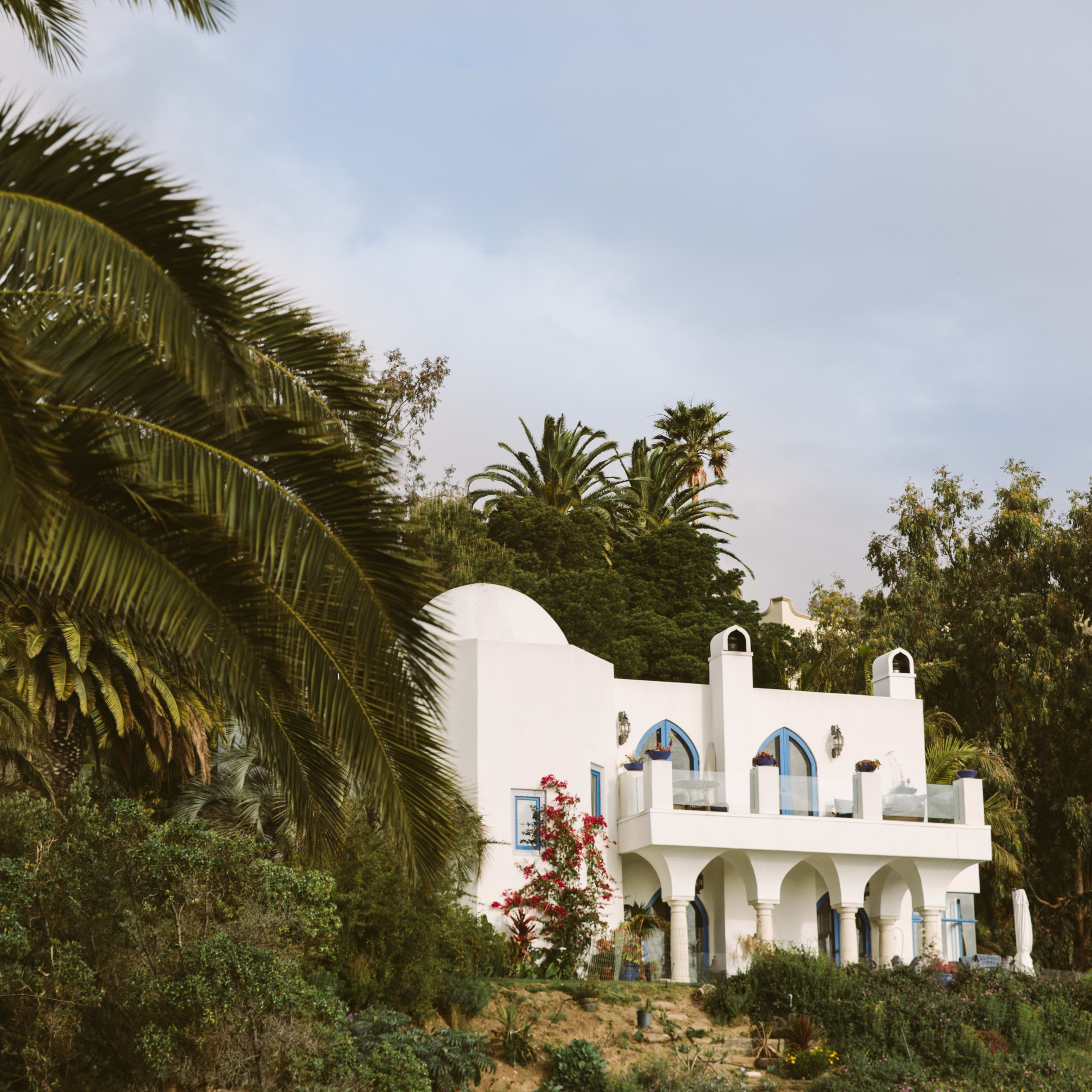 A white building with arched windows and palm trees surrounds it, situated on a hillside.
