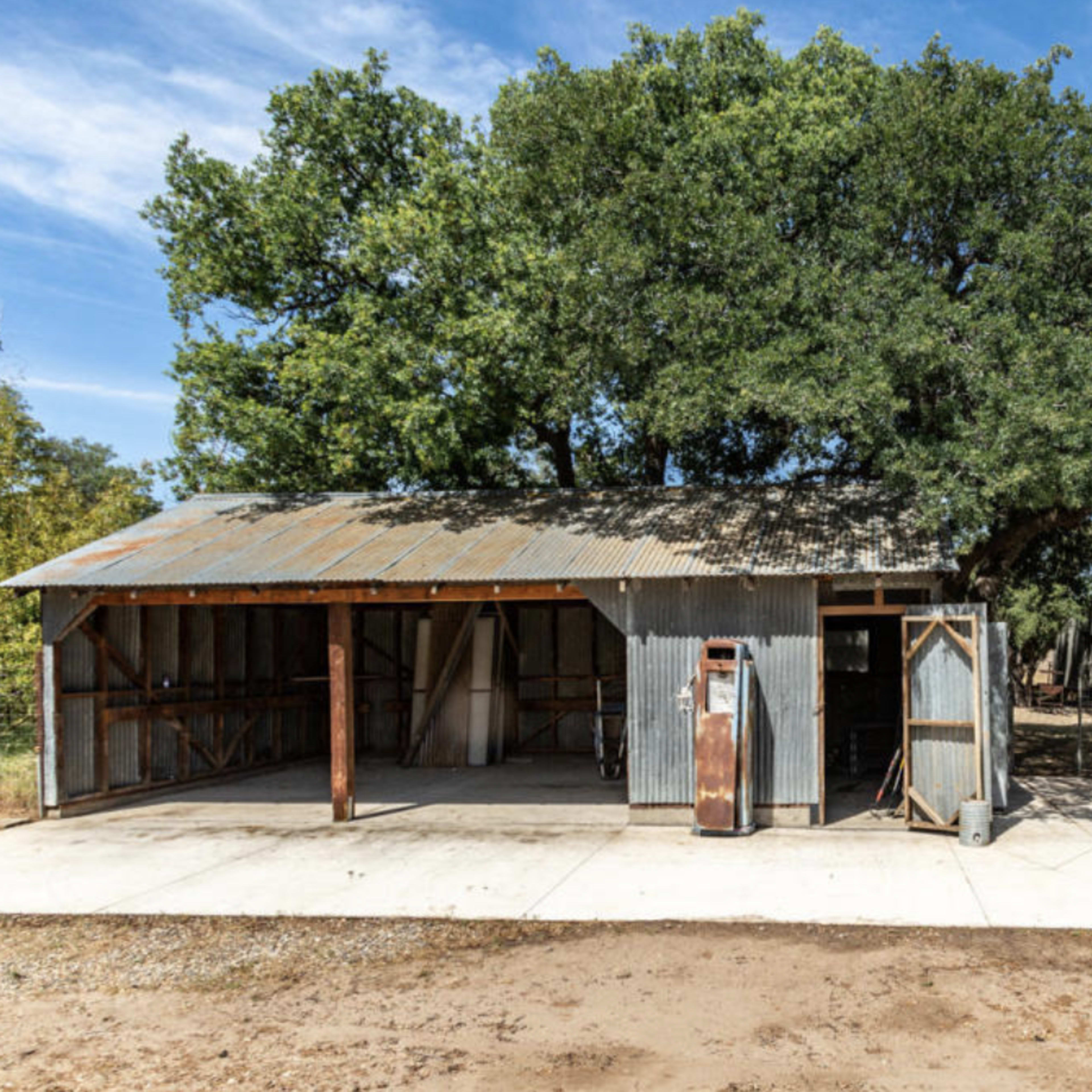A weathered metal barn stands beside a large oak tree, with a concrete pad in front and a nearby fenced area.