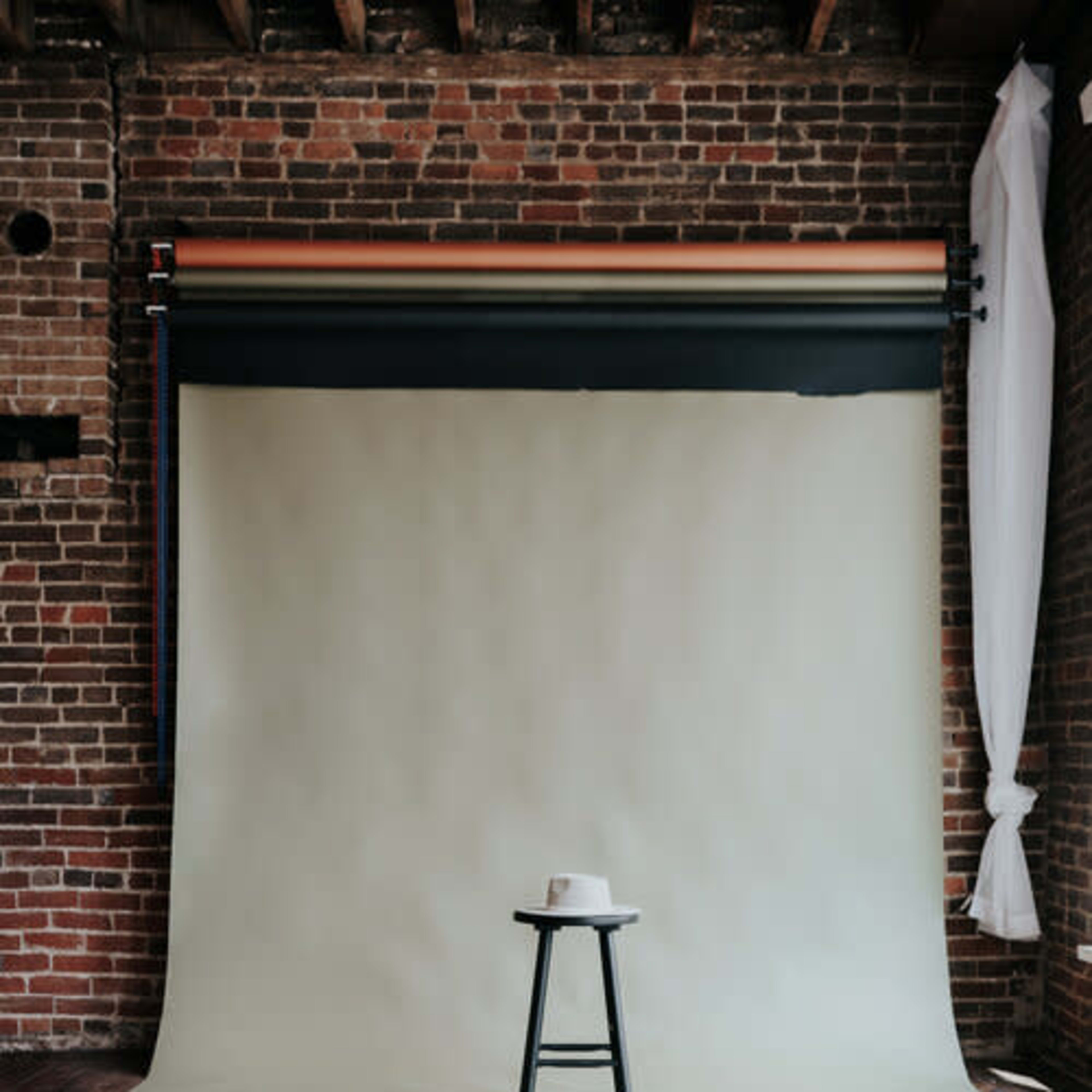 A simple black stool with a white hat sits in front of a pale backdrop and brick wall in a studio setting.