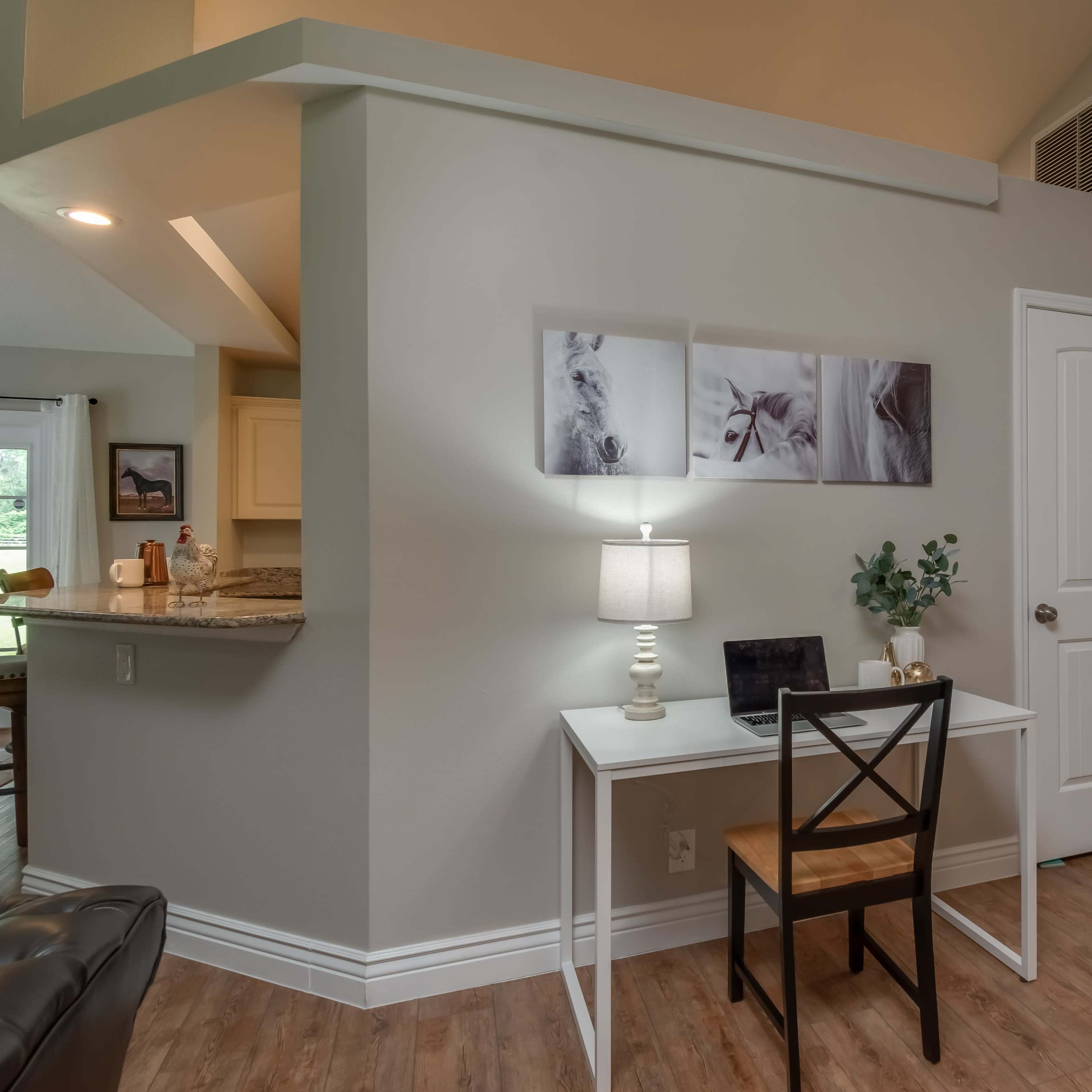 A small study area with a white desk and chair next to a wall adorned with framed photographs, leading into a doorway and a kitchen area in the background.