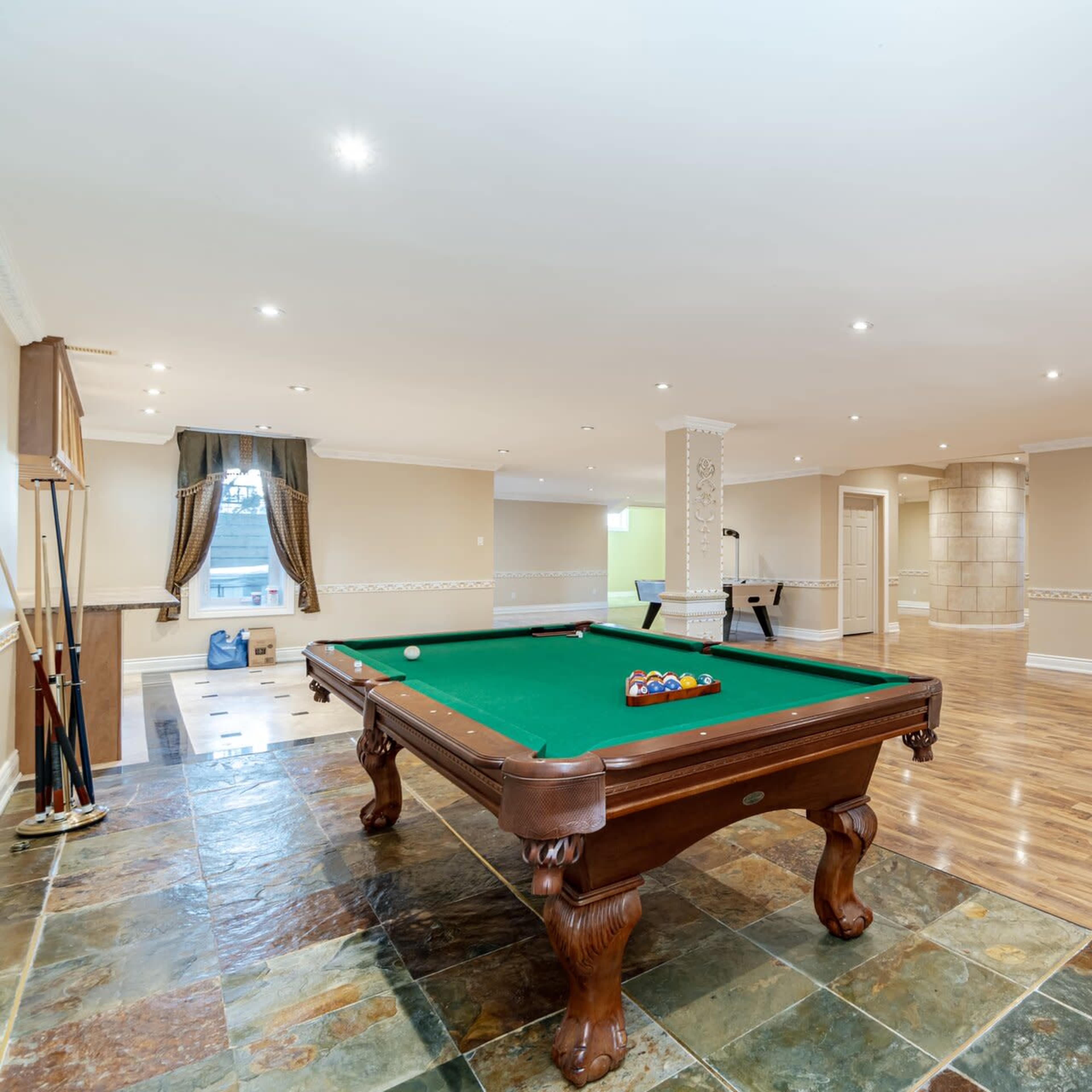 A spacious game room featuring a green billiard table, polished wooden flooring, and well-lit areas with smooth walls.