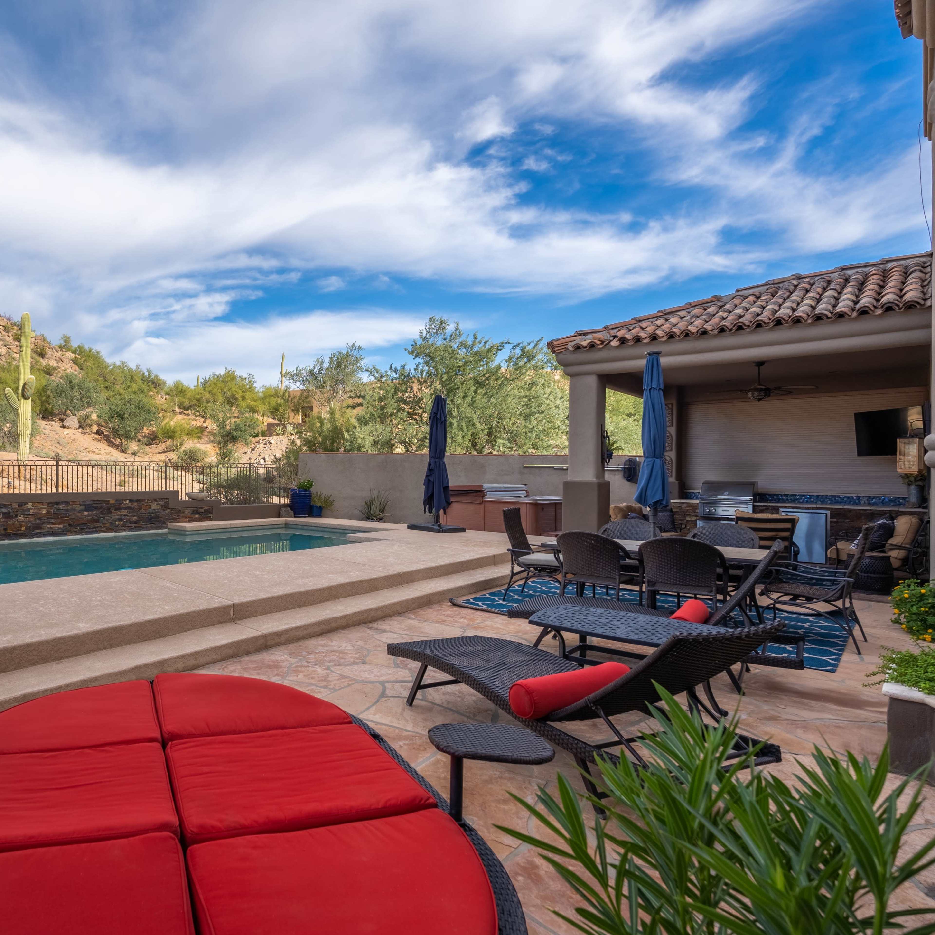 A landscaped backyard features a pool surrounded by lounge chairs and a covered seating area, with a desert landscape and mountains in the background.