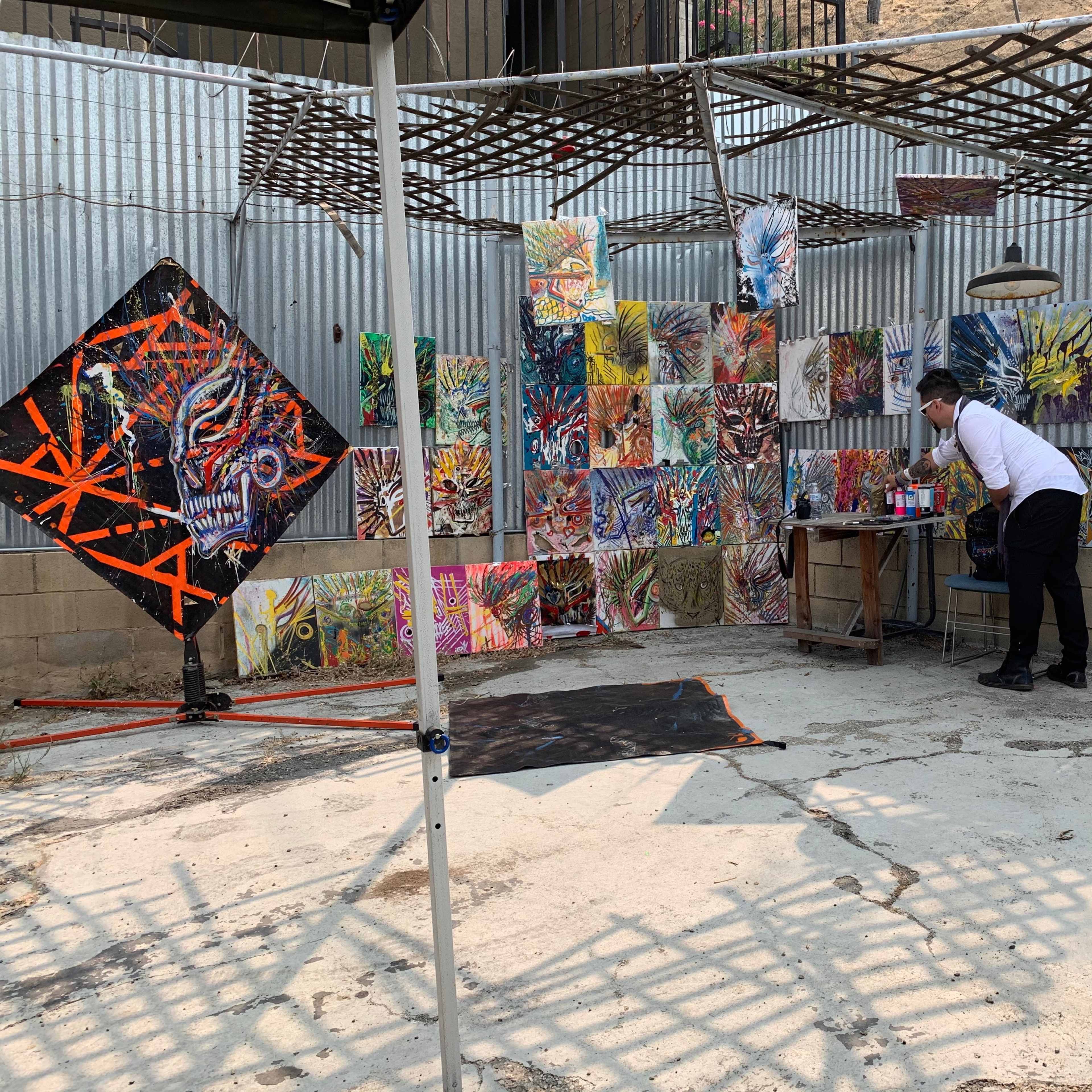 An artist is working on a table in a creative outdoor space surrounded by colorful paintings displayed on the walls.