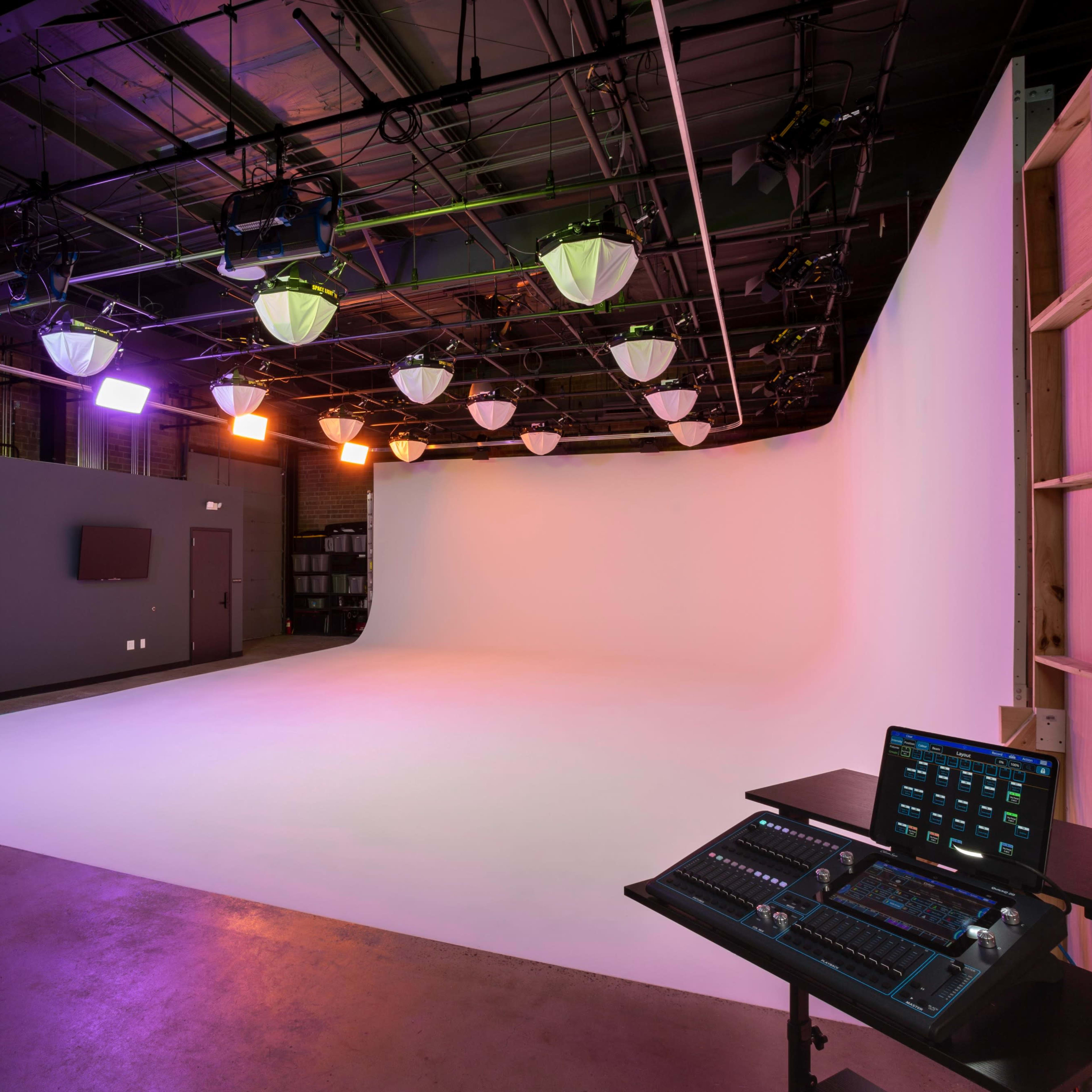 A modern studio with a white backdrop, green curtain, and lighting fixtures, along with a control panel on the side.