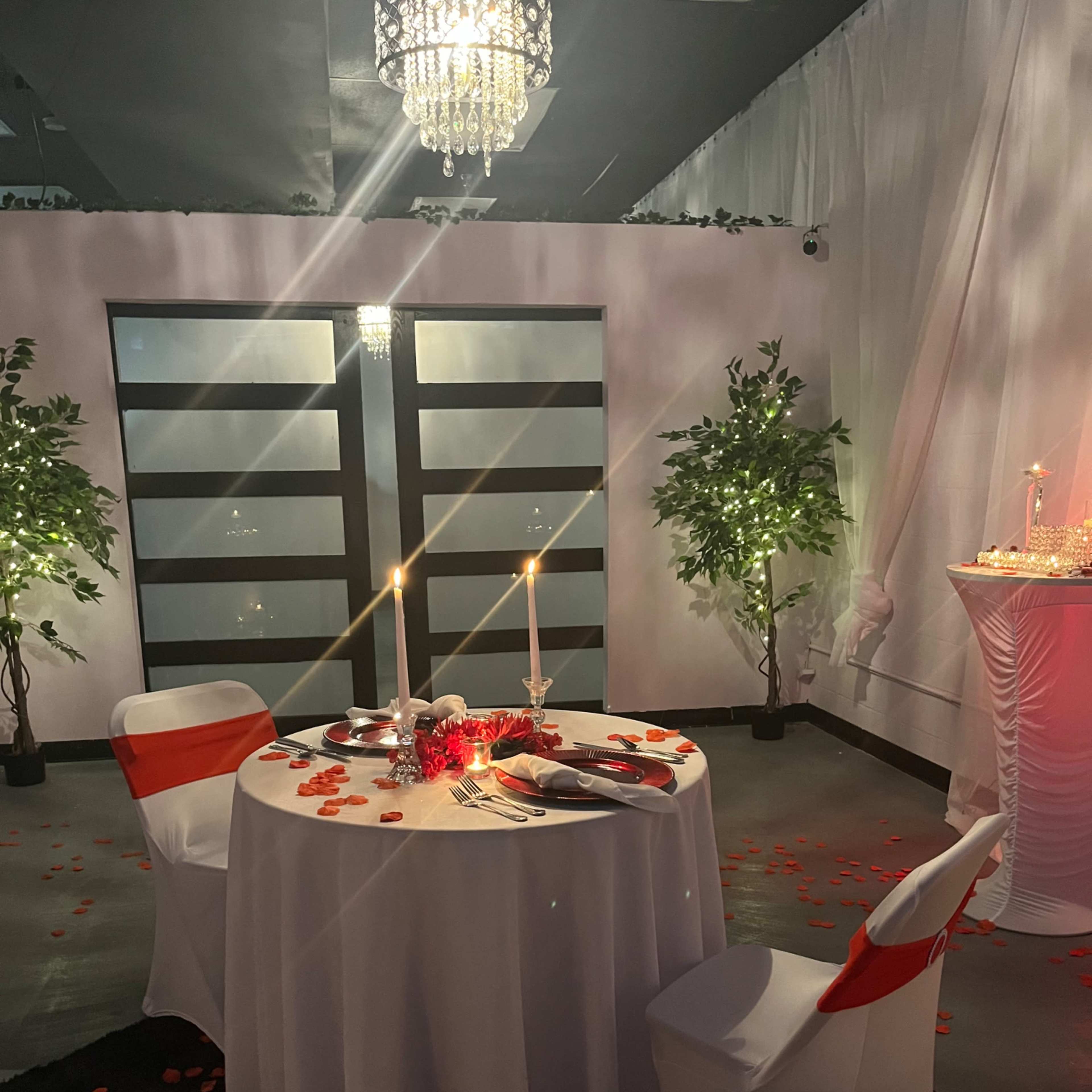 A romantic dinner setup features a round table with a white tablecloth, two chairs adorned with orange sashes, candles, rose petals, and a backdrop of two dark doors.