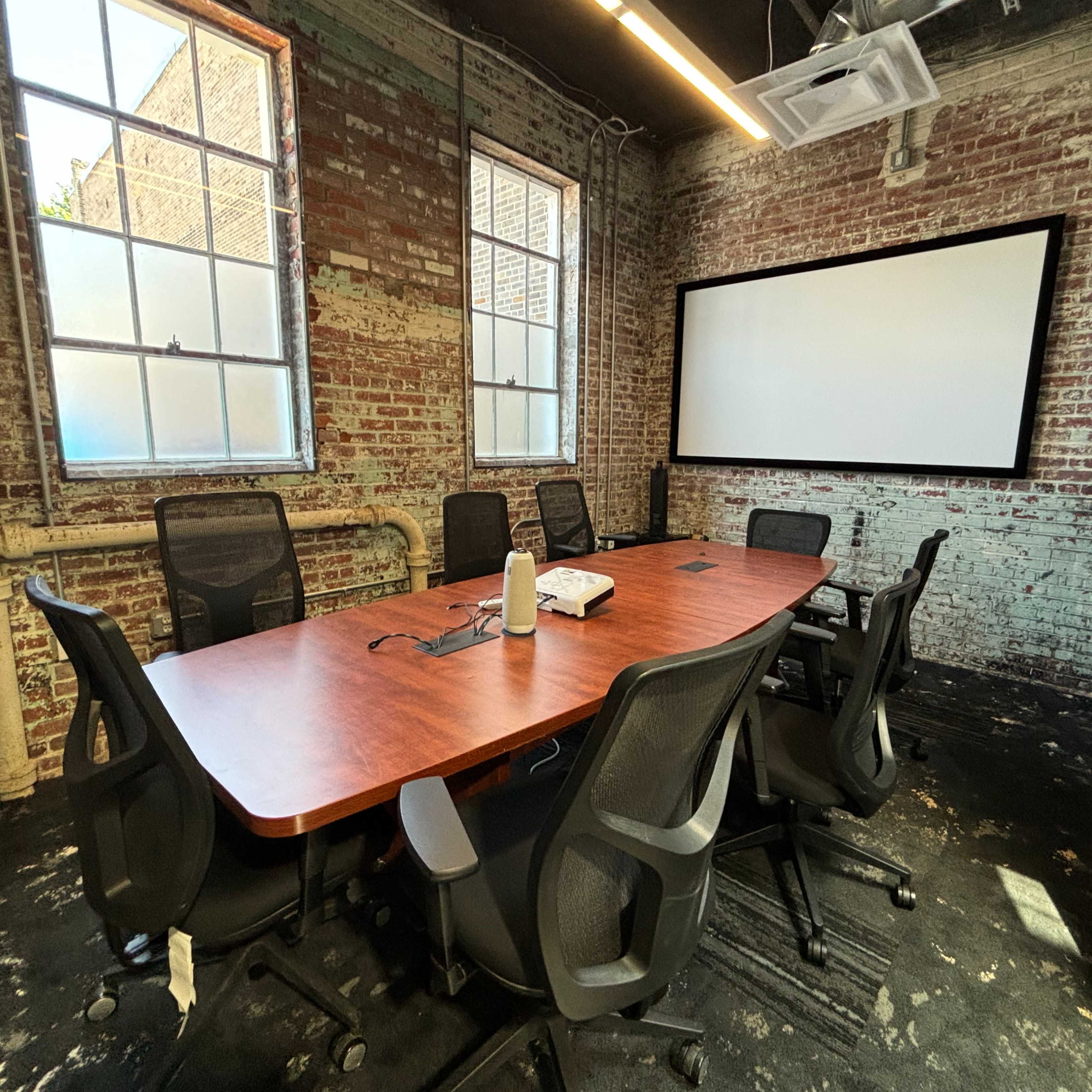 A conference room features a long wooden table surrounded by black chairs, with exposed brick walls and a large screen on one side.
