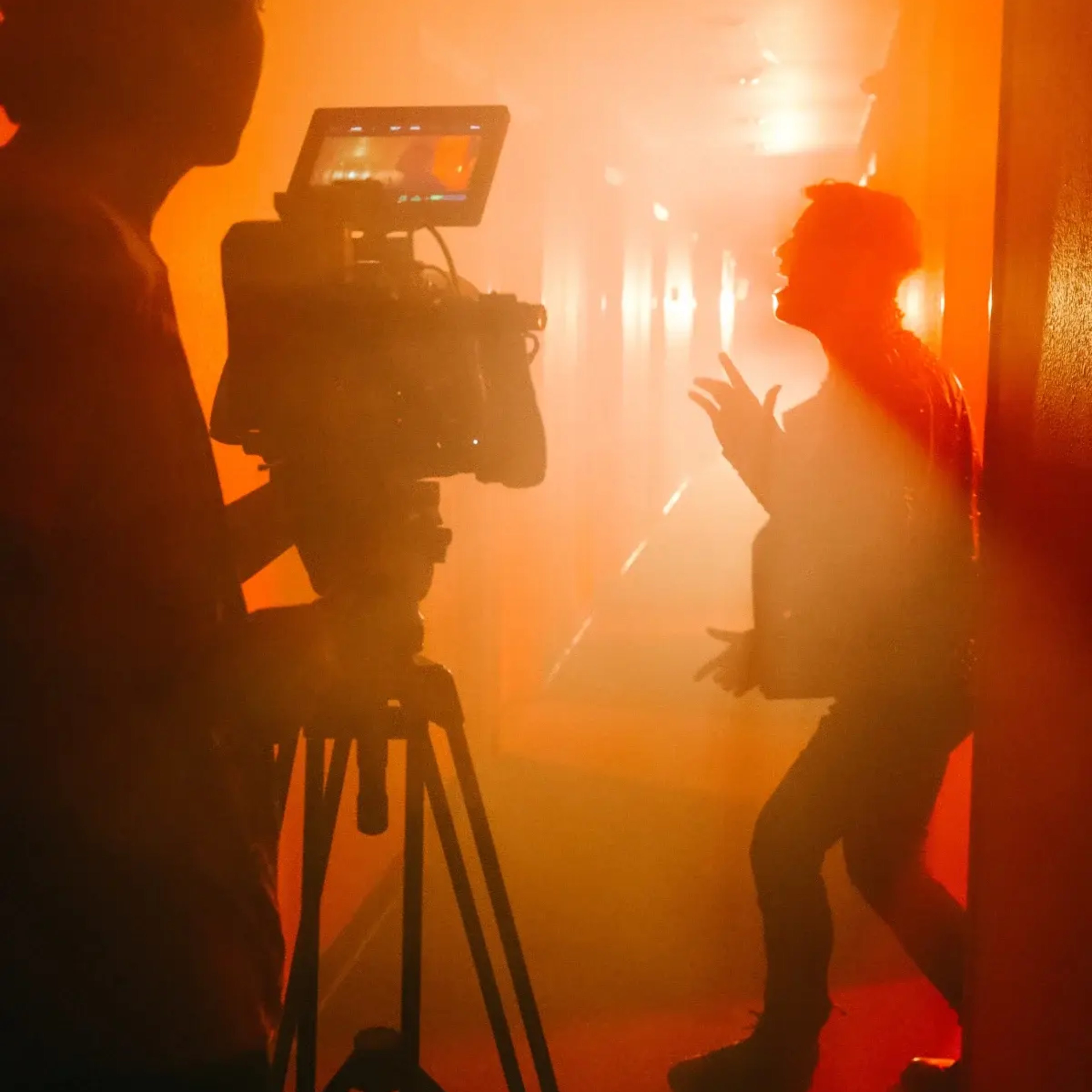 A person films another individual performing in a foggy hallway illuminated by orange light.