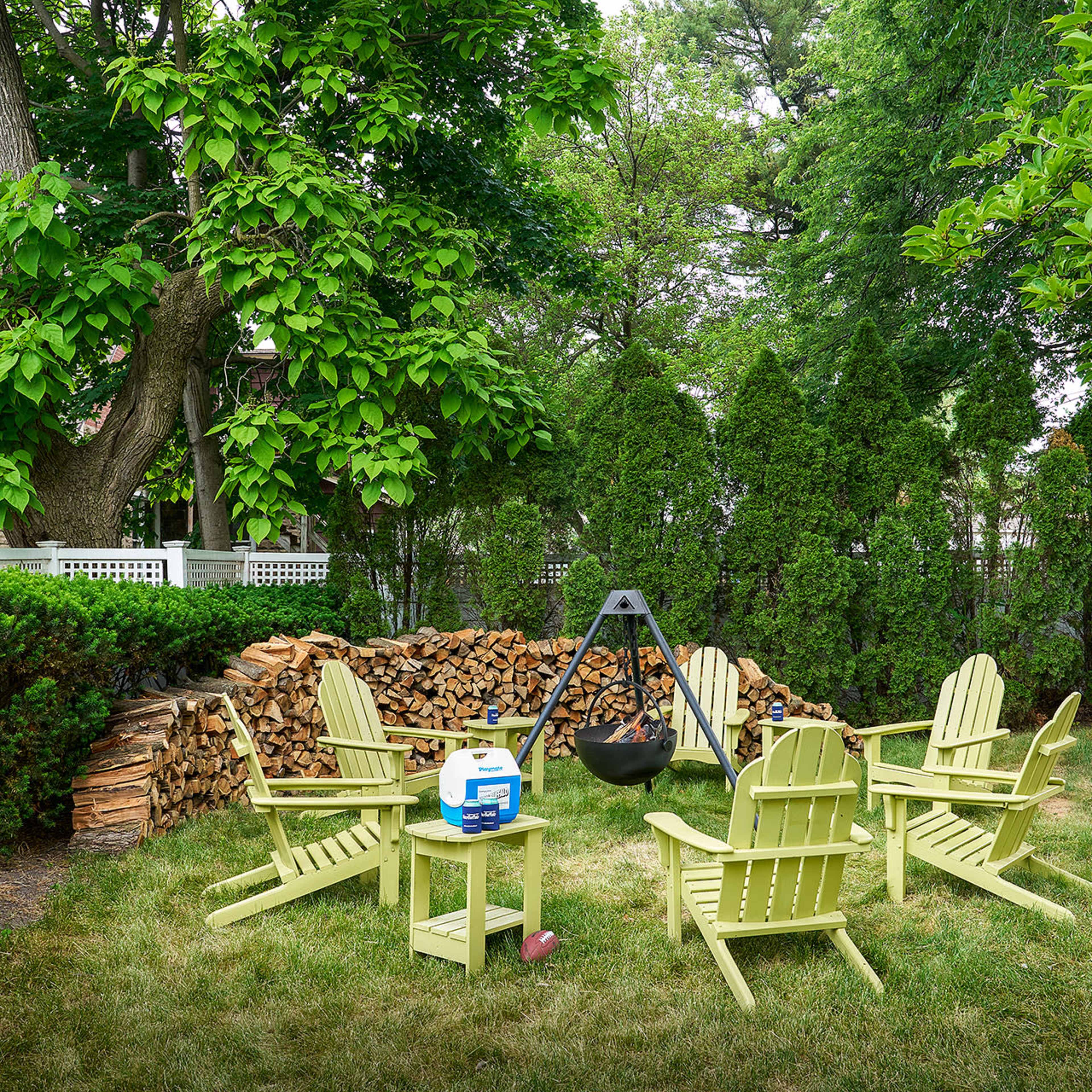 A circular arrangement of light green Adirondack chairs surrounds a fire pit in a grassy yard, with a stack of firewood and tall hedges in the background.