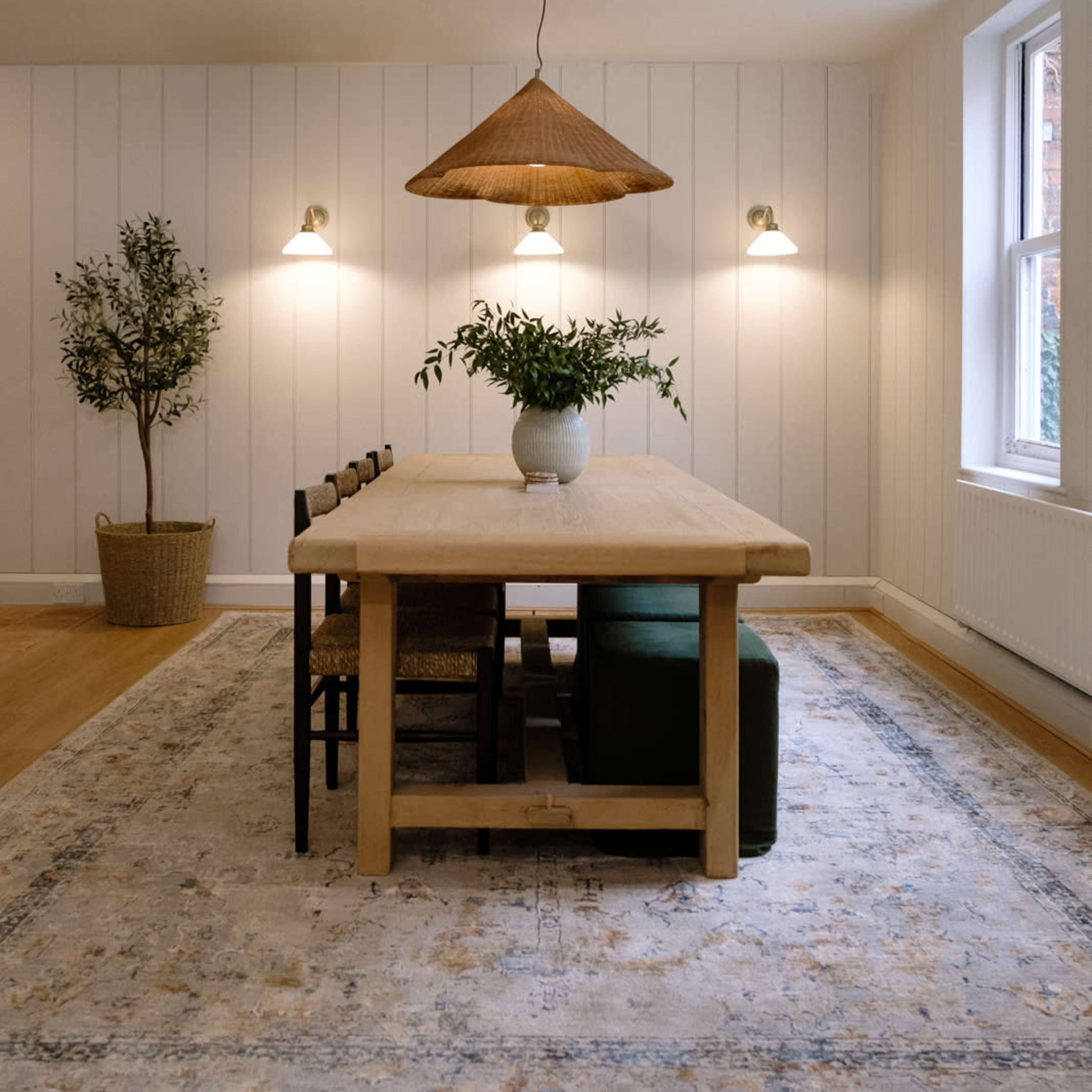 A wooden dining table is set in a softly lit room featuring white walls, green seating, and a hanging pendant light with a woven shade.
