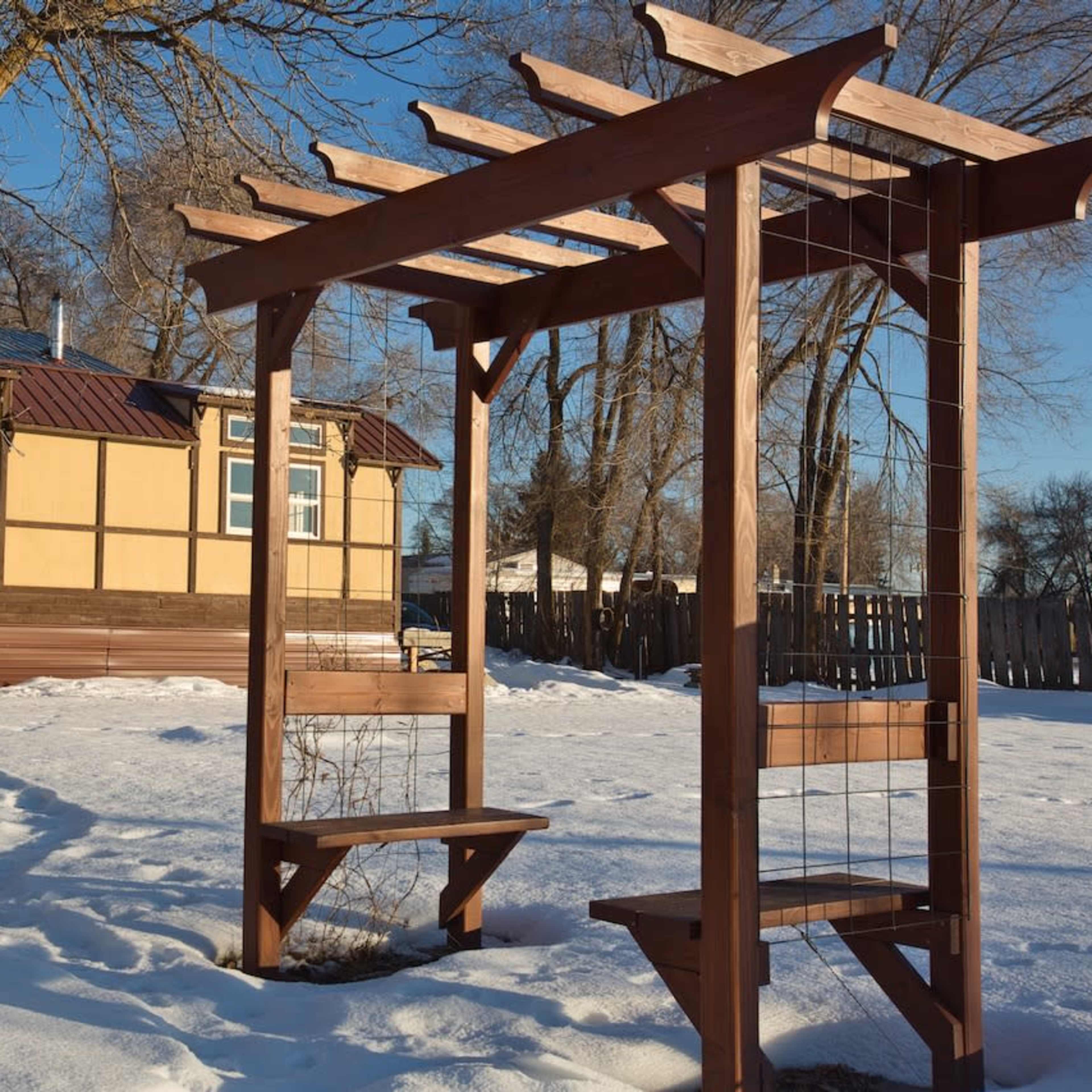 A wooden pergola stands in a snowy yard, with a house and stacked logs visible in the background.