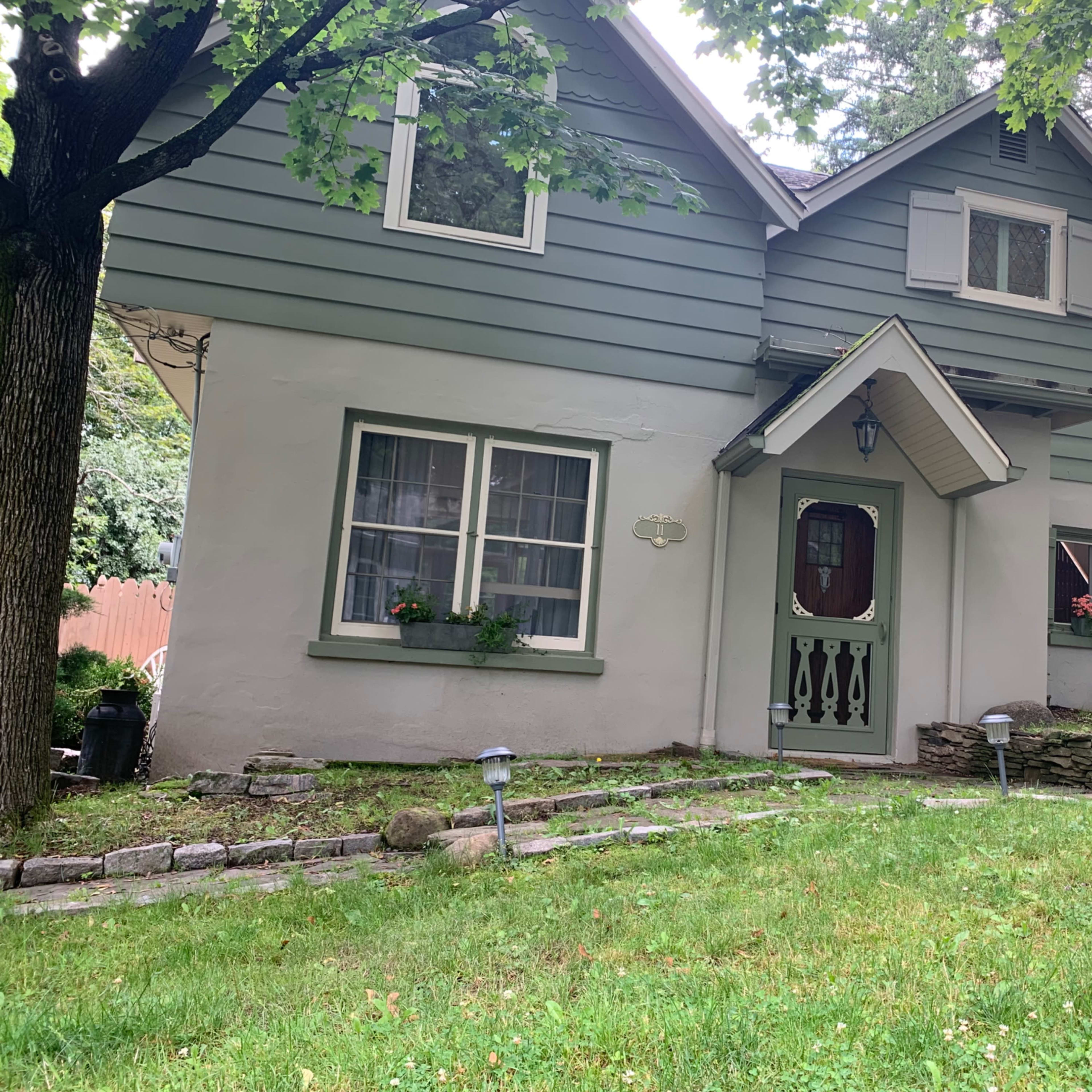 A two-story house with a green and white exterior, surrounded by grass and trees, and featuring a small stone pathway leading to the entrance.