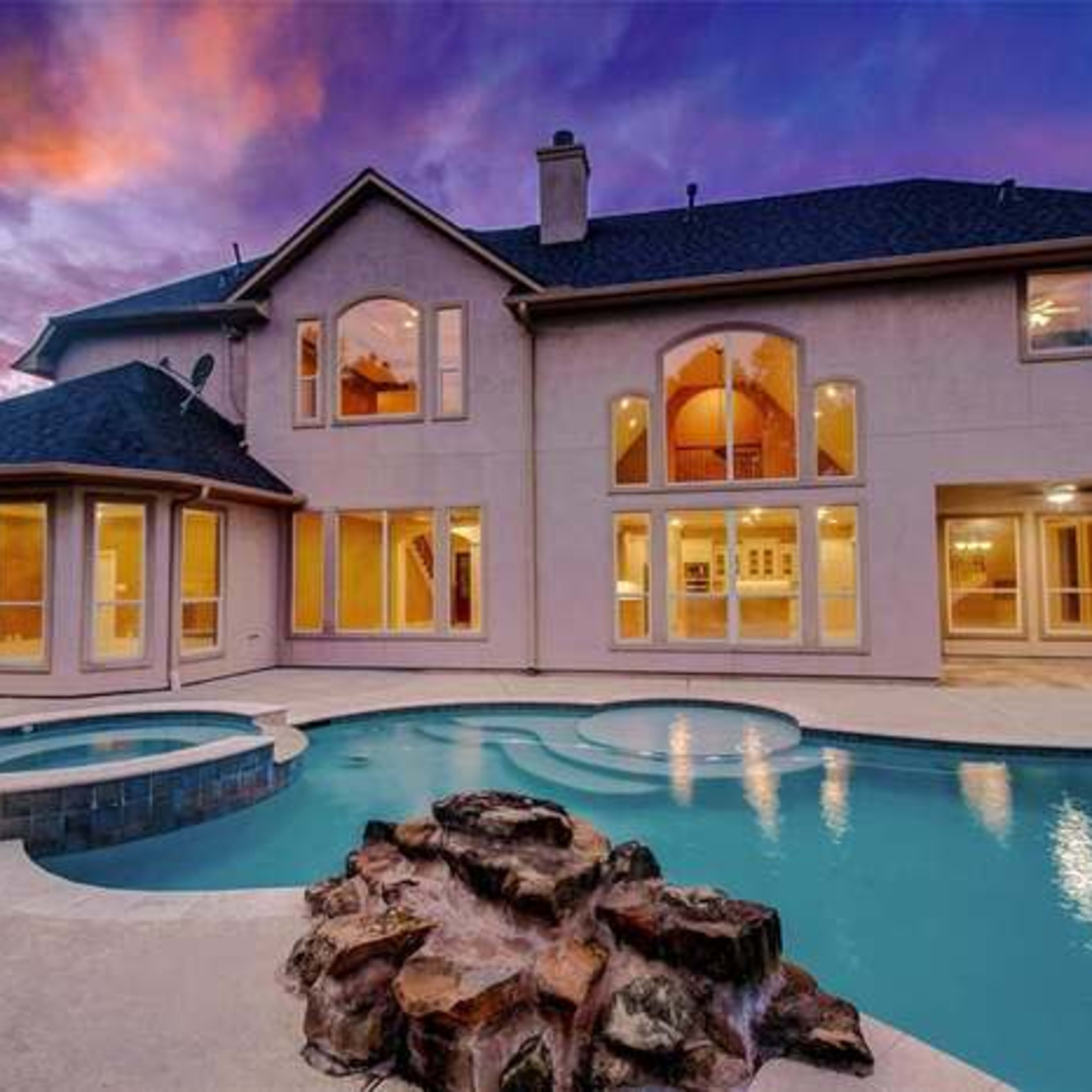 A modern two-story house with large windows, a swimming pool with a spa, and a landscaped backyard against a vibrant sunset sky.