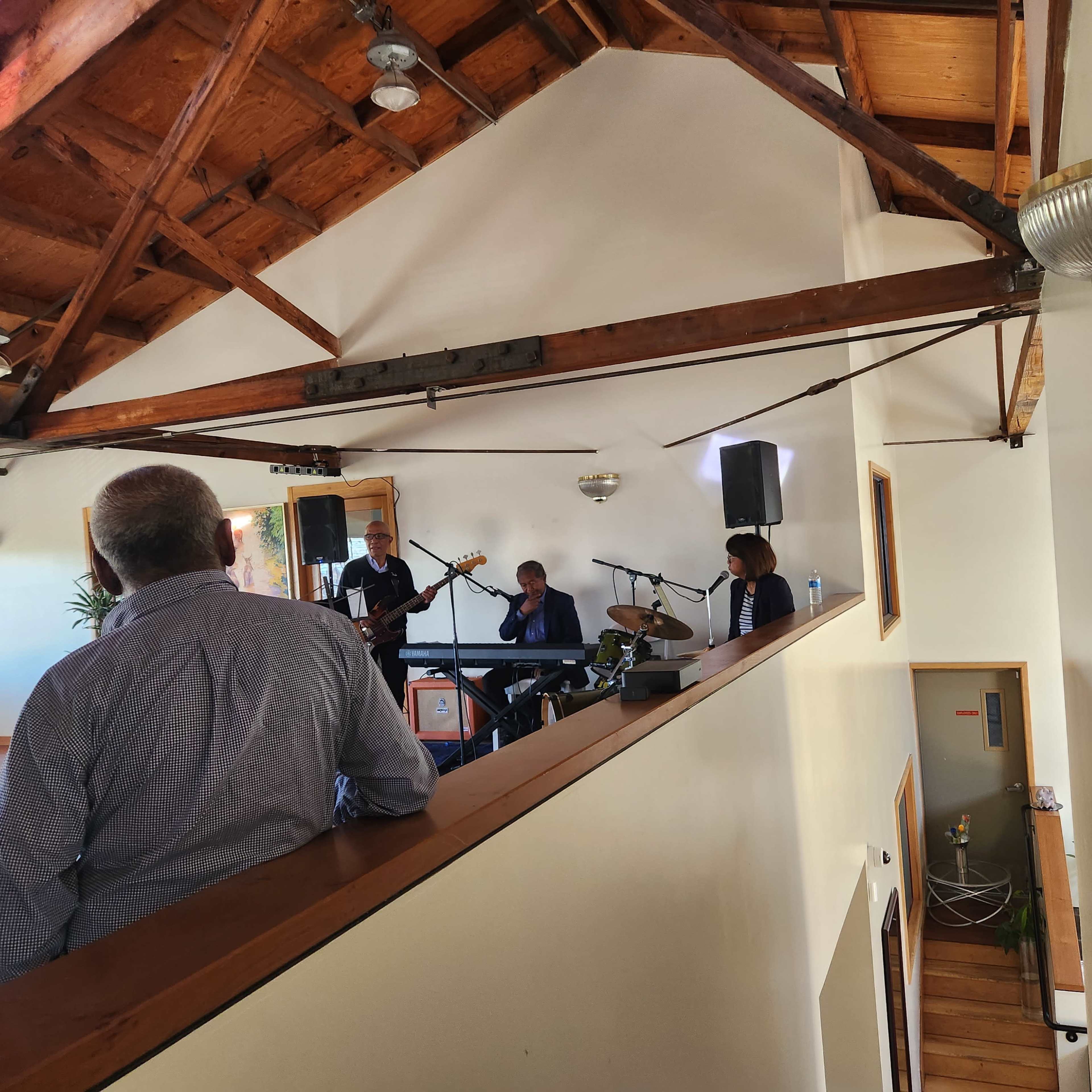 A small musical performance is taking place in a loft-style venue, with musicians on stage and an audience member looking on from a balcony.