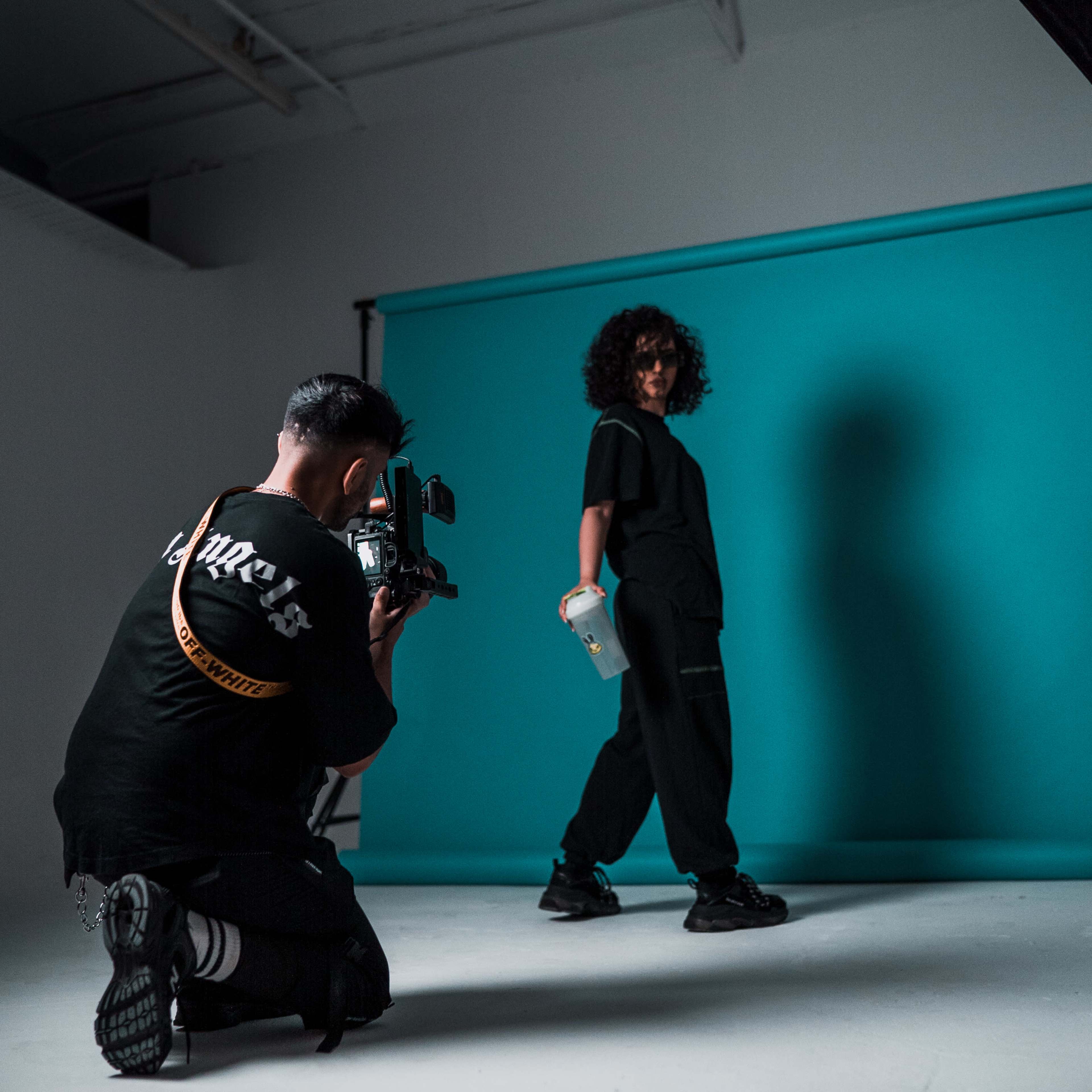 A photographer captures a model holding a water bottle against a turquoise backdrop in a studio setting.
