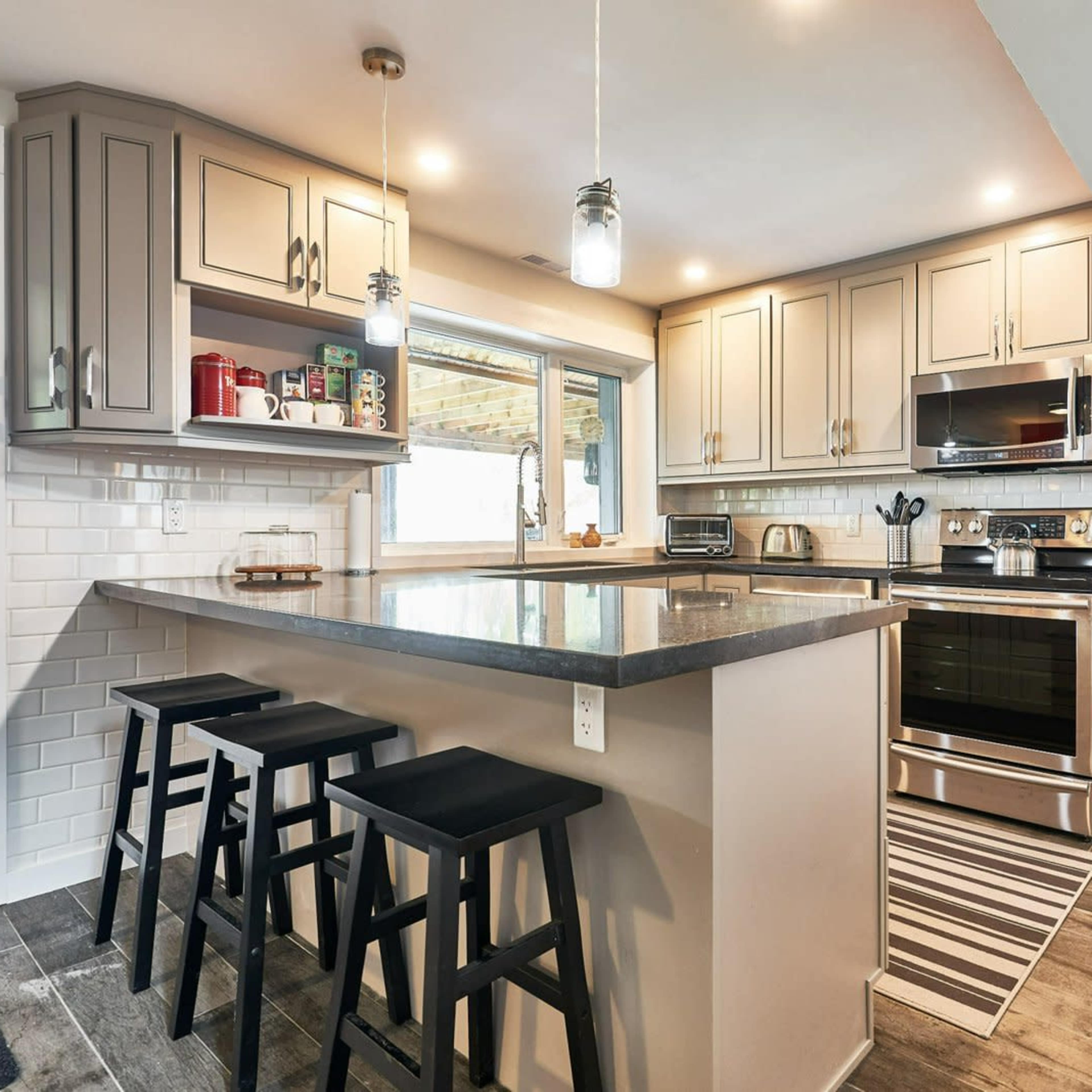A modern kitchen featuring a central island with three black stools, stainless steel appliances, and light gray cabinets.