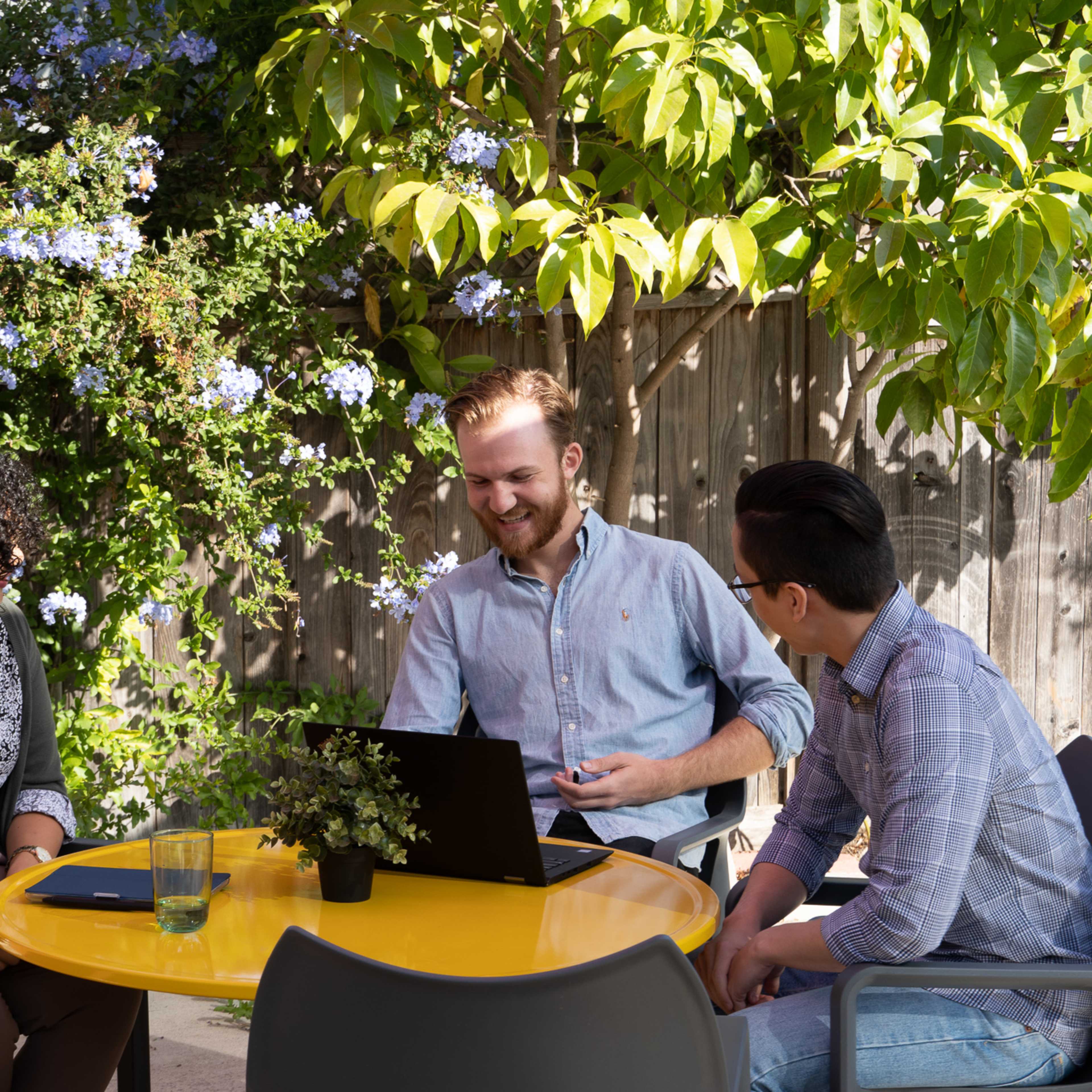 Three people sit at a yellow table in a garden, engaged in conversation with a laptop and plants around them.
