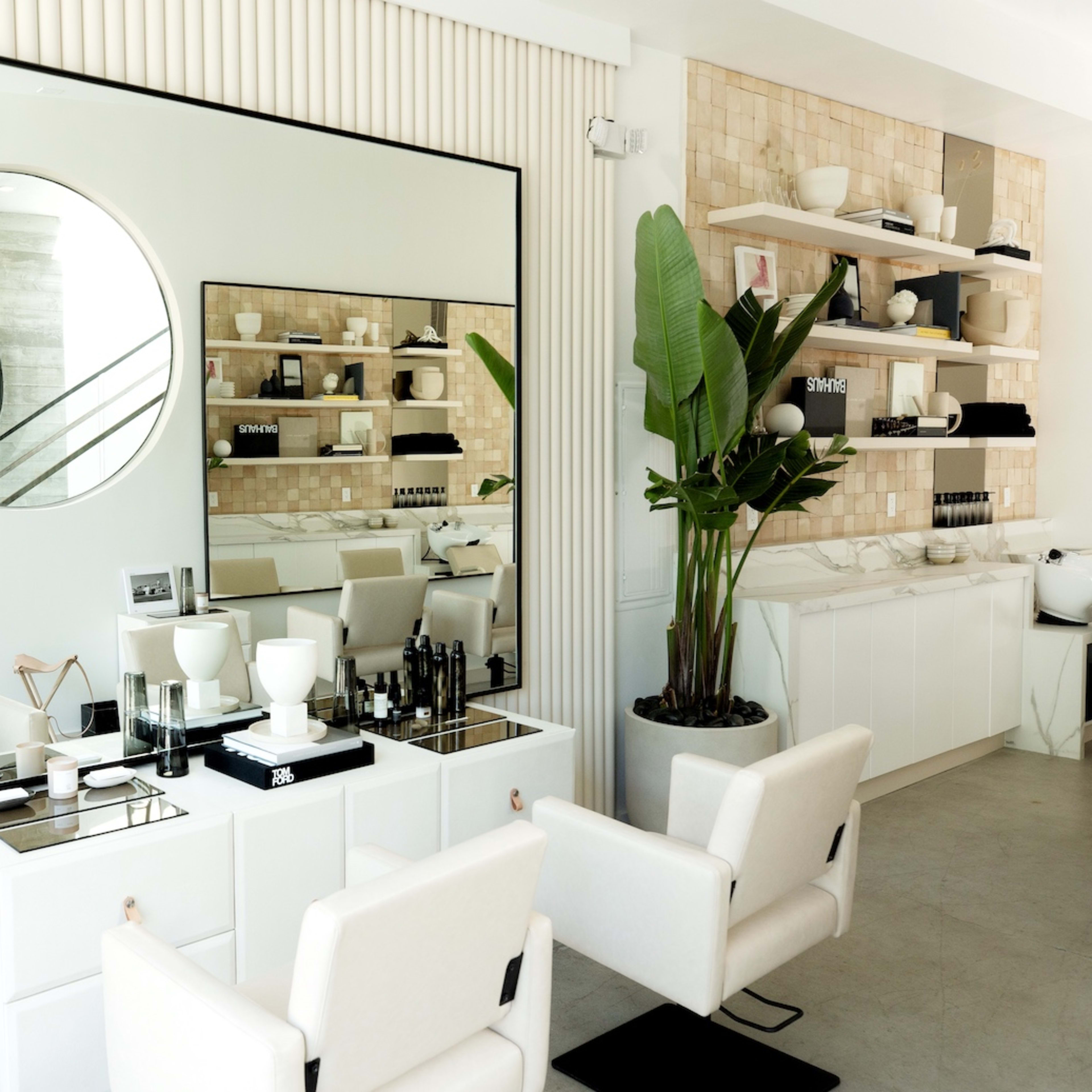 A modern hair salon with a large circular mirror, minimalist furniture, and neatly arranged products on a shelf in a bright, open space.