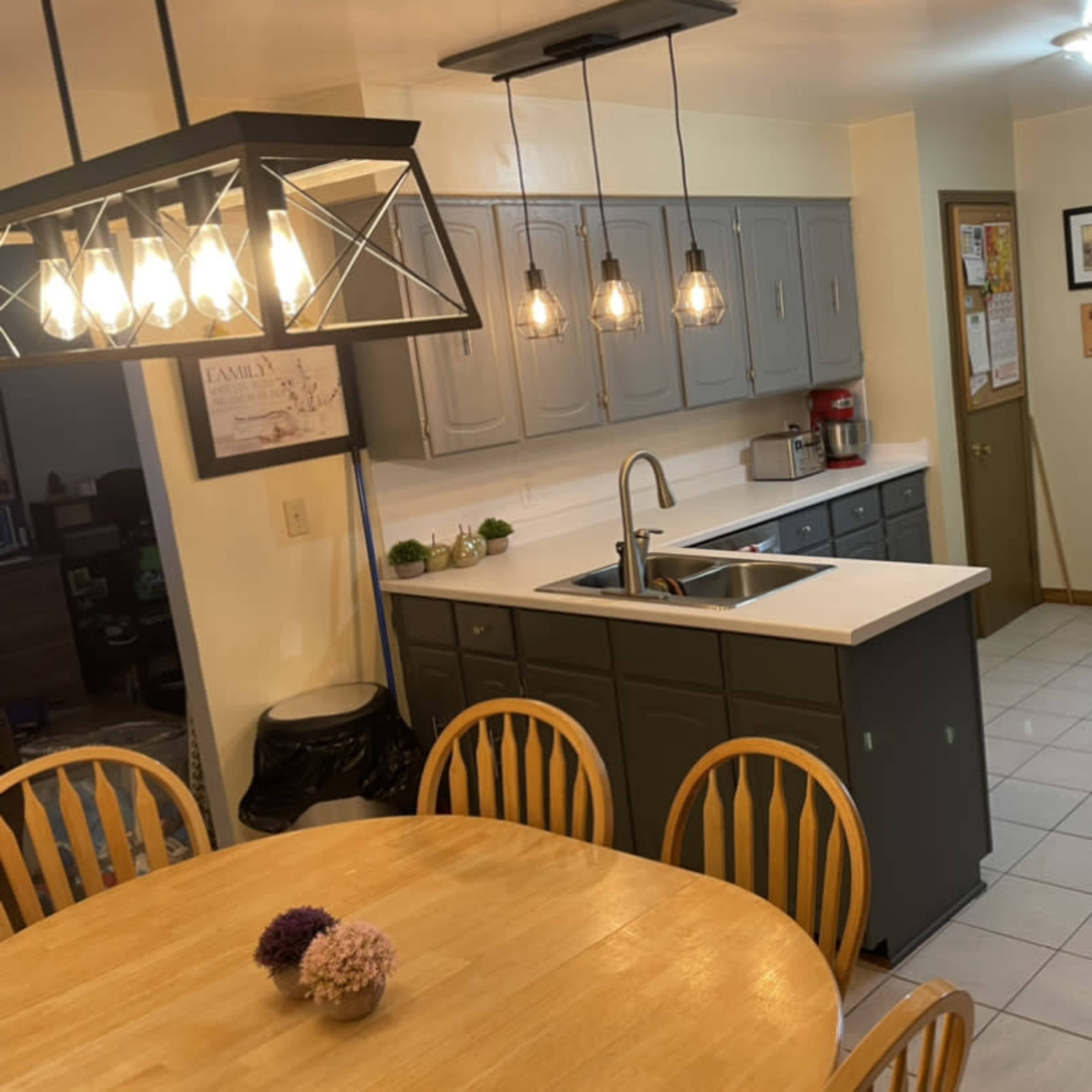 A bright kitchen with gray cabinets, a central island with a sink, a wooden dining table, and a doorway leading to another room.
