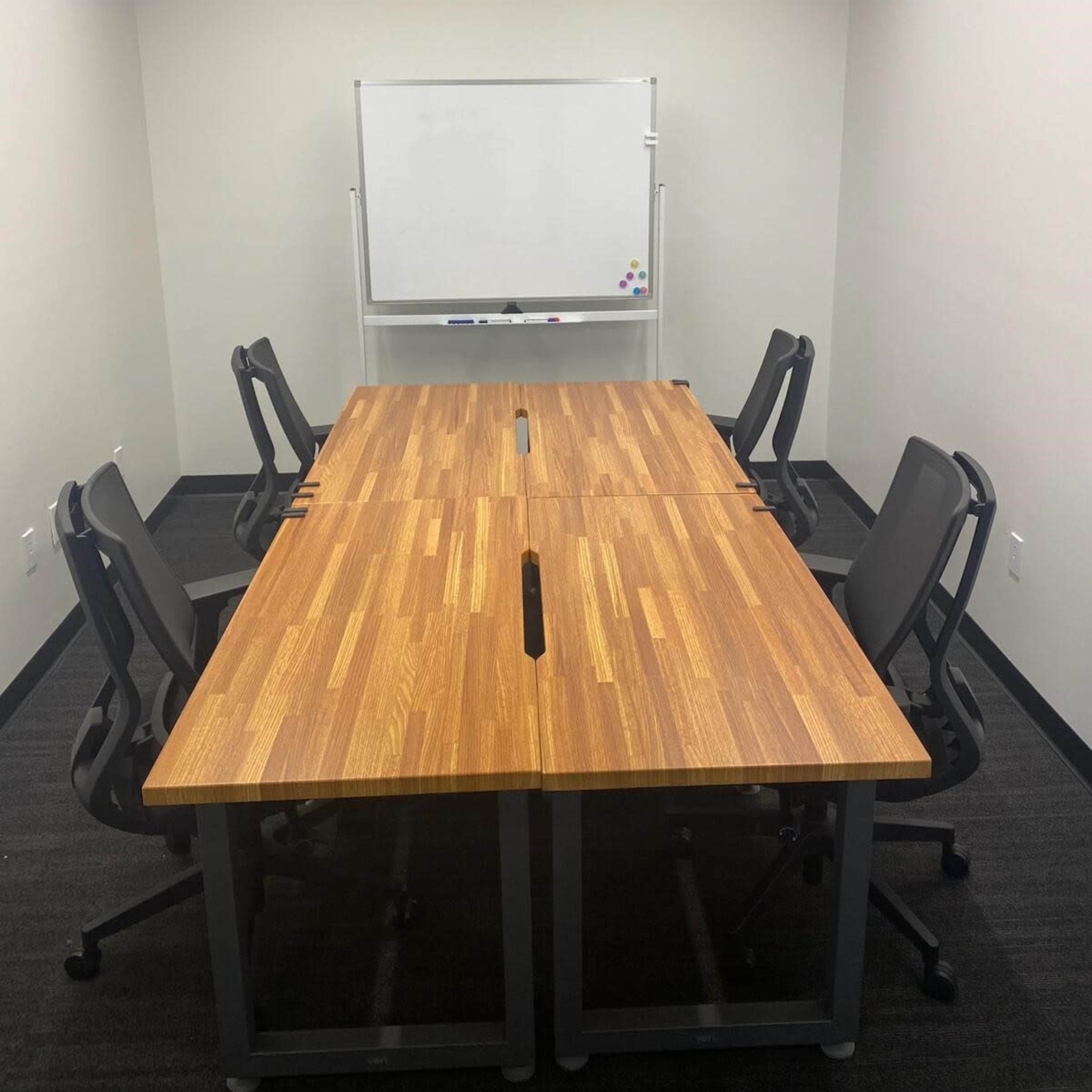 A conference room features a rectangular wooden table with six ergonomic chairs, accompanied by a whiteboard on one wall.