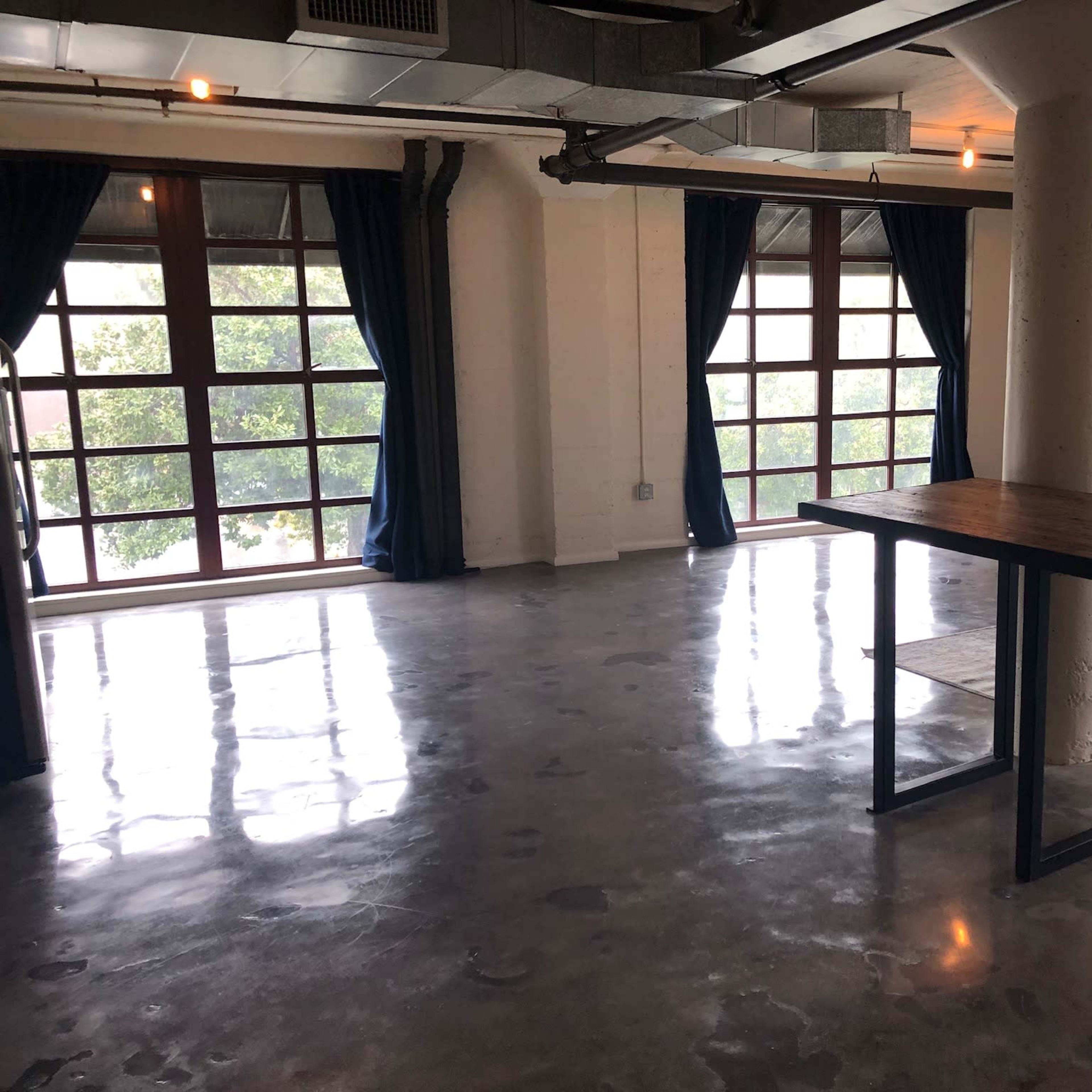 A spacious, empty room with polished concrete floors, large windows, and a wooden table in the center.